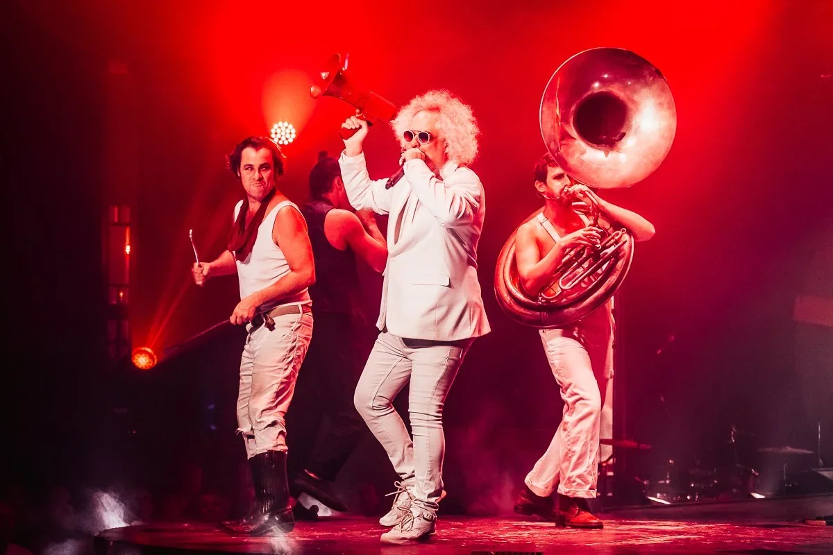 Musicians performing on stage with red lighting, including a man with curly blonde hair singing into a microphone, a man with a drumstick, and a person playing a sousaphone.