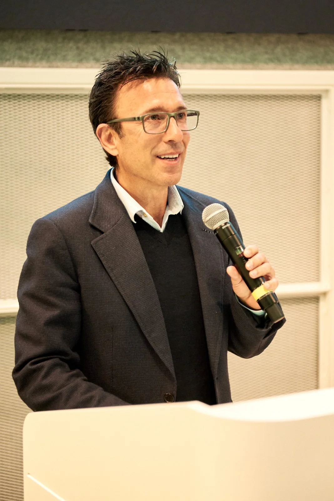A man in glasses and a dark blazer speaking into a microphone in front of a gray textured background.