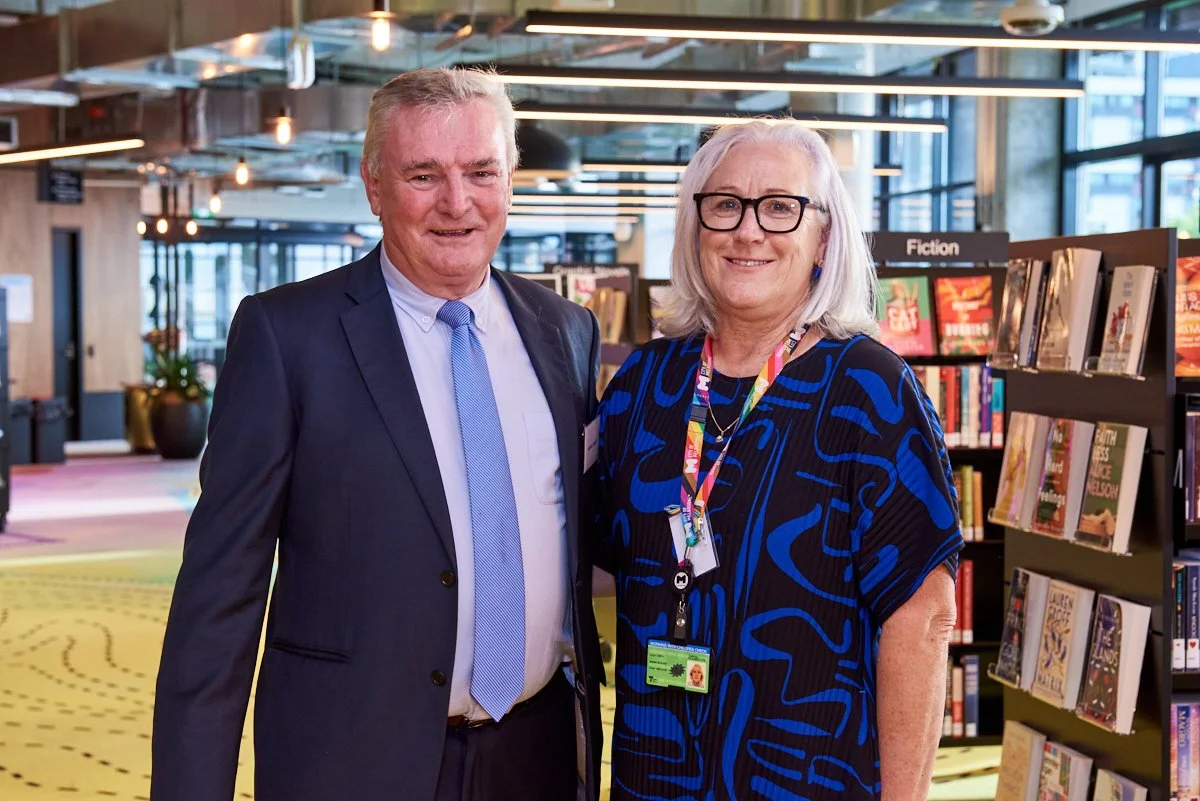 A man and a woman standing together in a bookstore. The man is wearing a navy suit with a light blue tie, and the woman has white hair, glasses, and is wearing a blue and black patterned dress. Both are smiling.