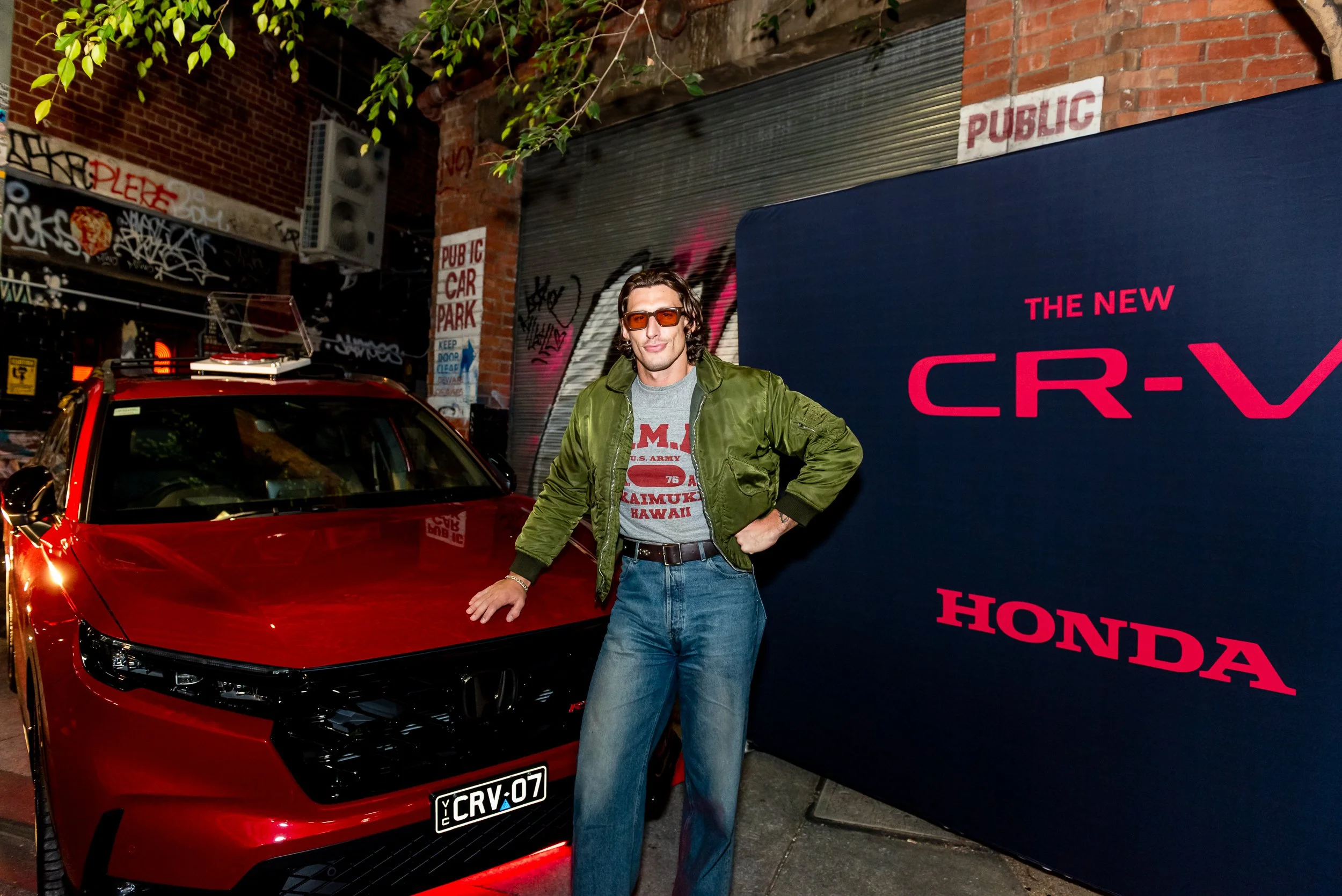 A man with long dark hair, wearing sunglasses, a gray t-shirt, and an olive green jacket, standing next to a red sports car with a custom license plate reading 'CRY-07' in an urban setting with graffiti on brick walls and a large dark blue sign that 
