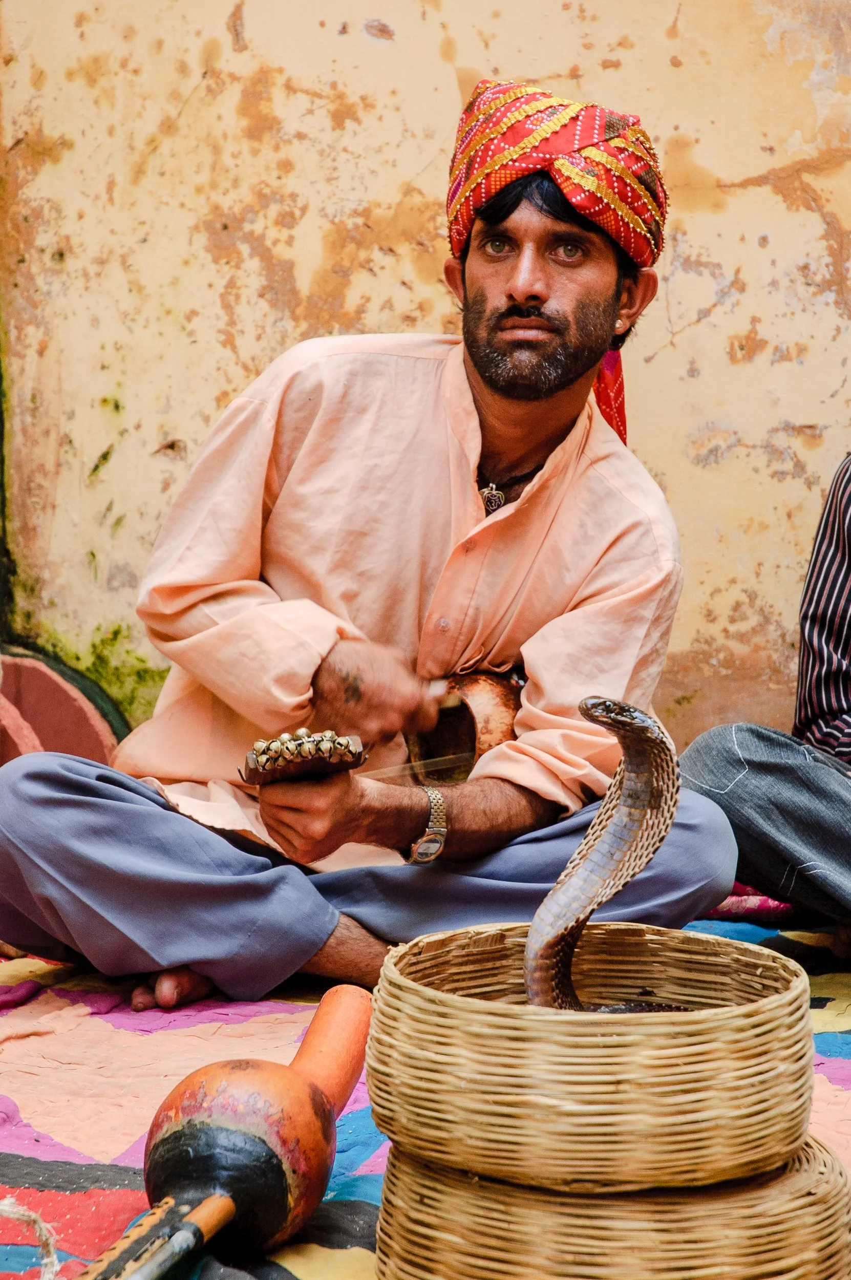 A man wearing traditional Indian clothing, including a colorful turban, sitting cross-legged on a mat. He is holding a cymbal in one hand and a javelin in the other. In front of him is a basket with a snake emerging from it, and a piece of bone or ca