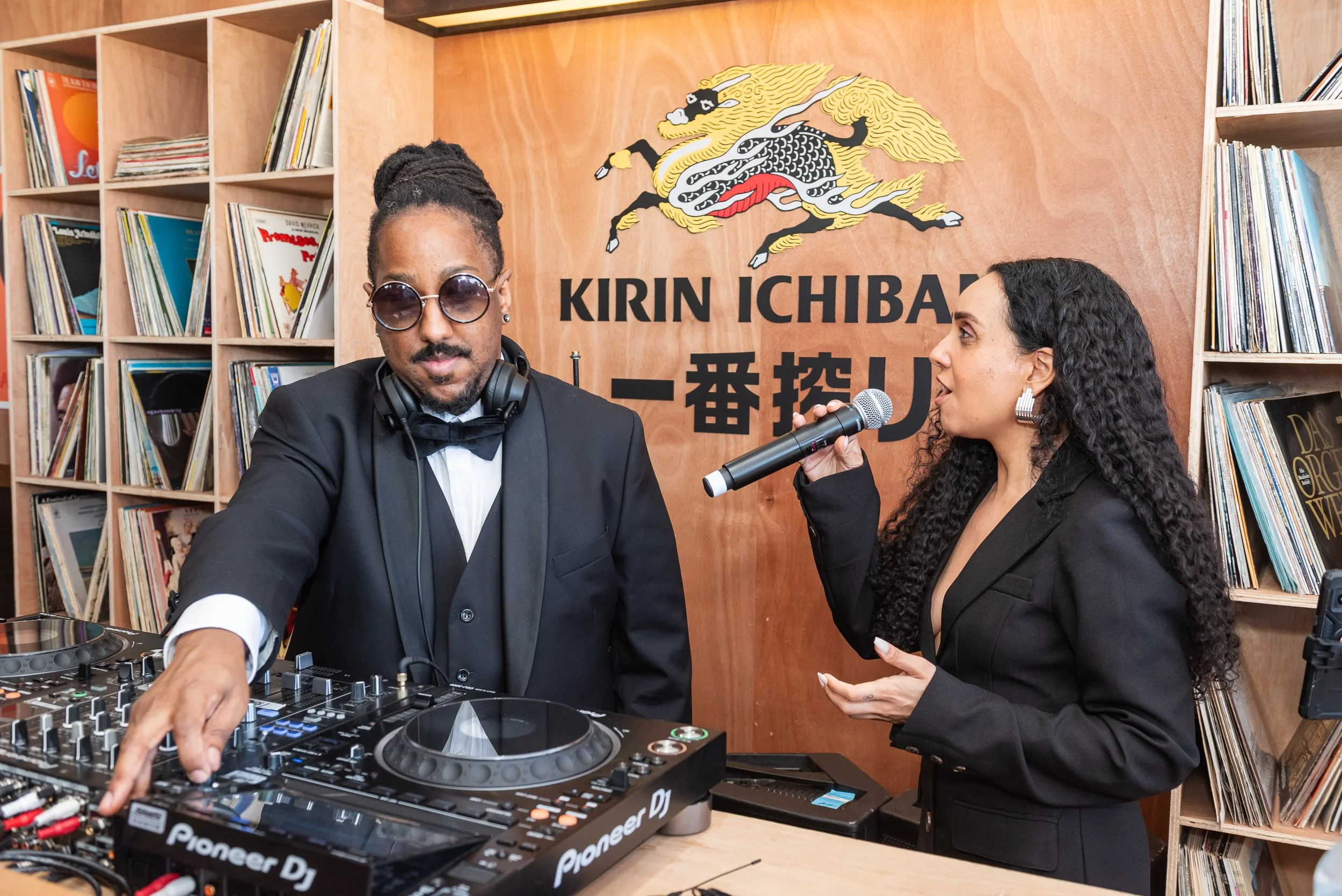 A DJ wearing sunglasses and headphones adjusting music equipment, while a woman with curly hair and earrings speaks into a microphone at a music event. They are inside a room with shelves of vinyl records and a sign that says "KIRIN ICHIBAN" with a d