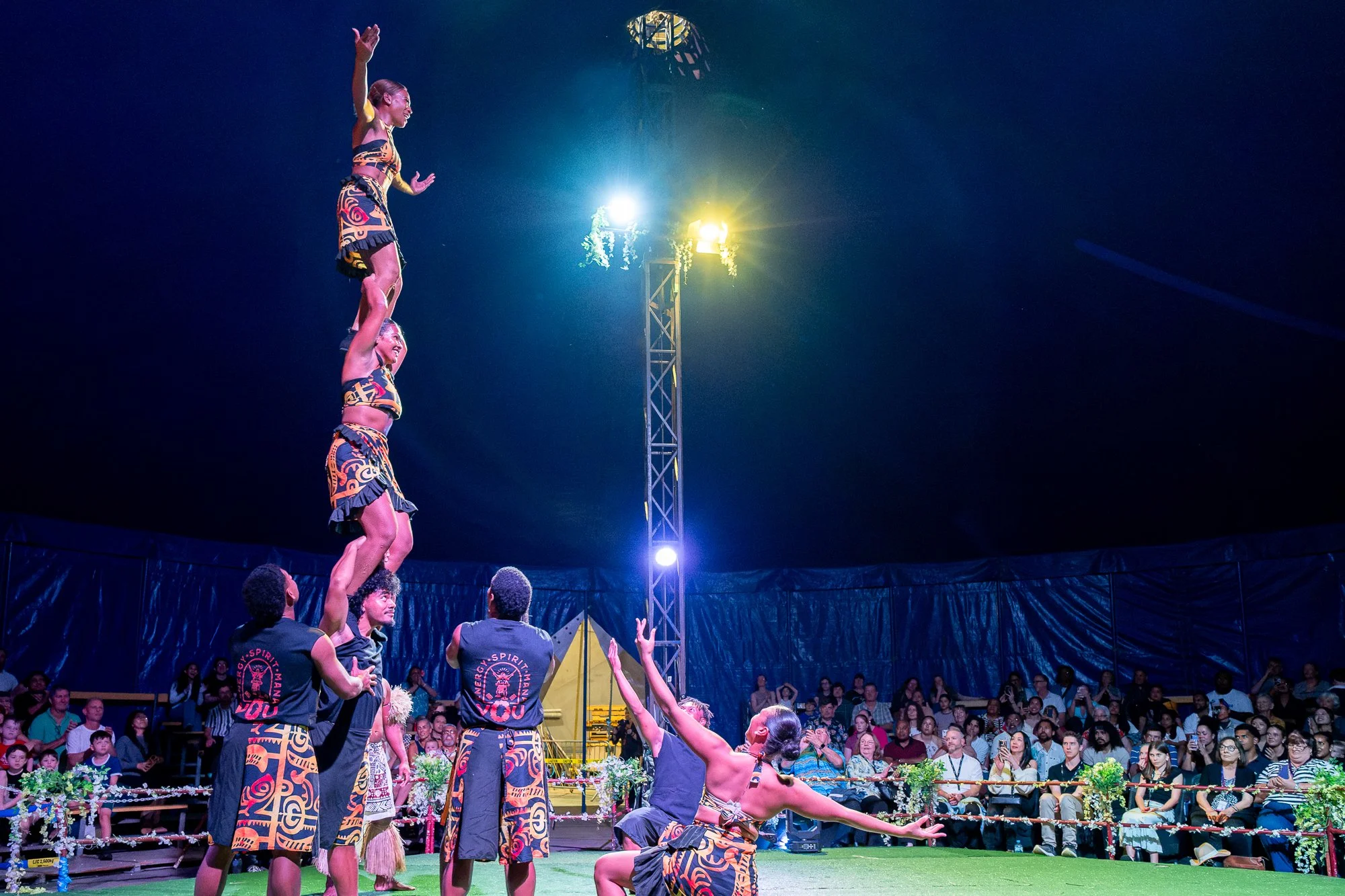 A performer standing on the hands of another performer in a circus act, surrounded by an audience watching the show at night.