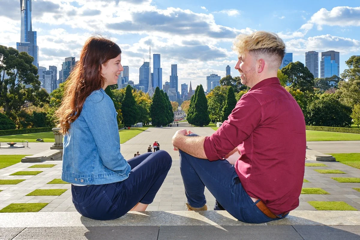 A young couple sitting on a stone ledge in a city park, facing each other and smiling, with a skyline of tall buildings and a partly cloudy sky in the background.