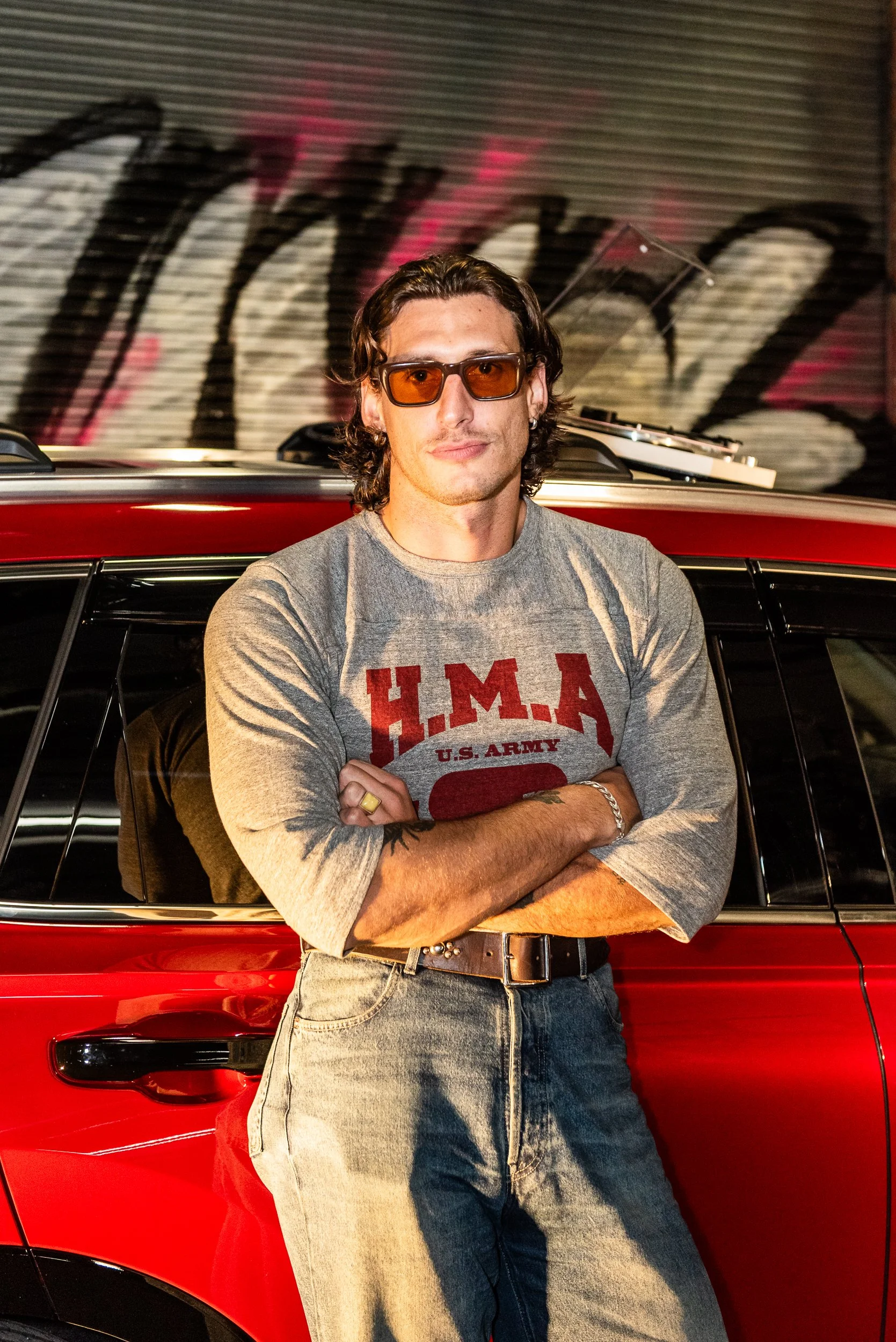 A man with wavy brown hair, wearing orange-tinted glasses, a gray long-sleeve shirt with red lettering, and high-waisted jeans, is standing with his arms crossed in front of a red car, in front of a graffiti-covered wall.