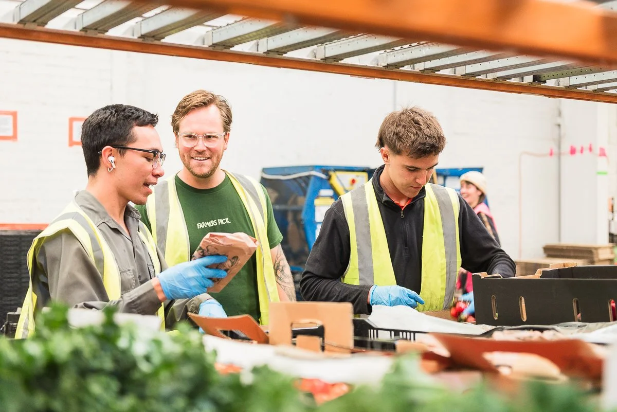 Three people working together in a warehouse or grocery store, wearing safety vests and gloves, with produce in front of them. Two men are looking at a clipboard, and a woman is sorting or packing items.
