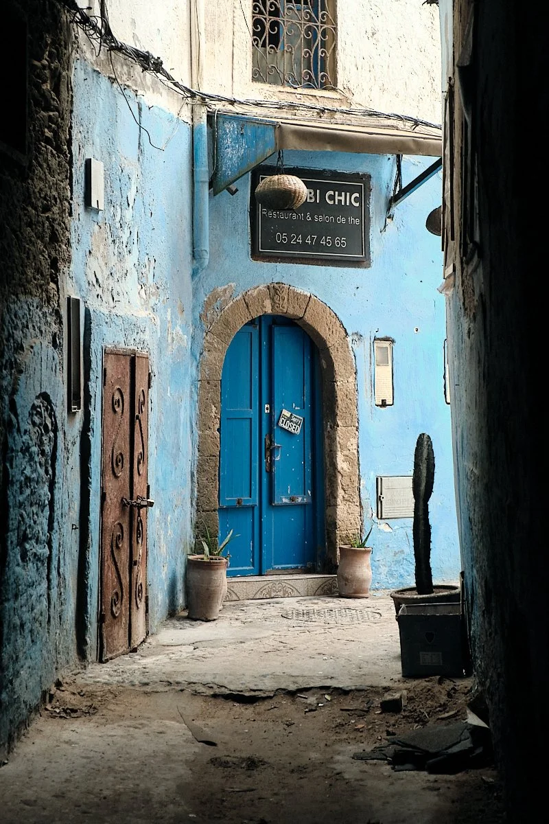 A narrow alley with a weathered blue door framed by a stone arch, two potted plants on either side, a cactus in a pot to the right, and a sign above the door indicating a restaurant and salon with a phone number. An 'Open' sign is crossed out and the