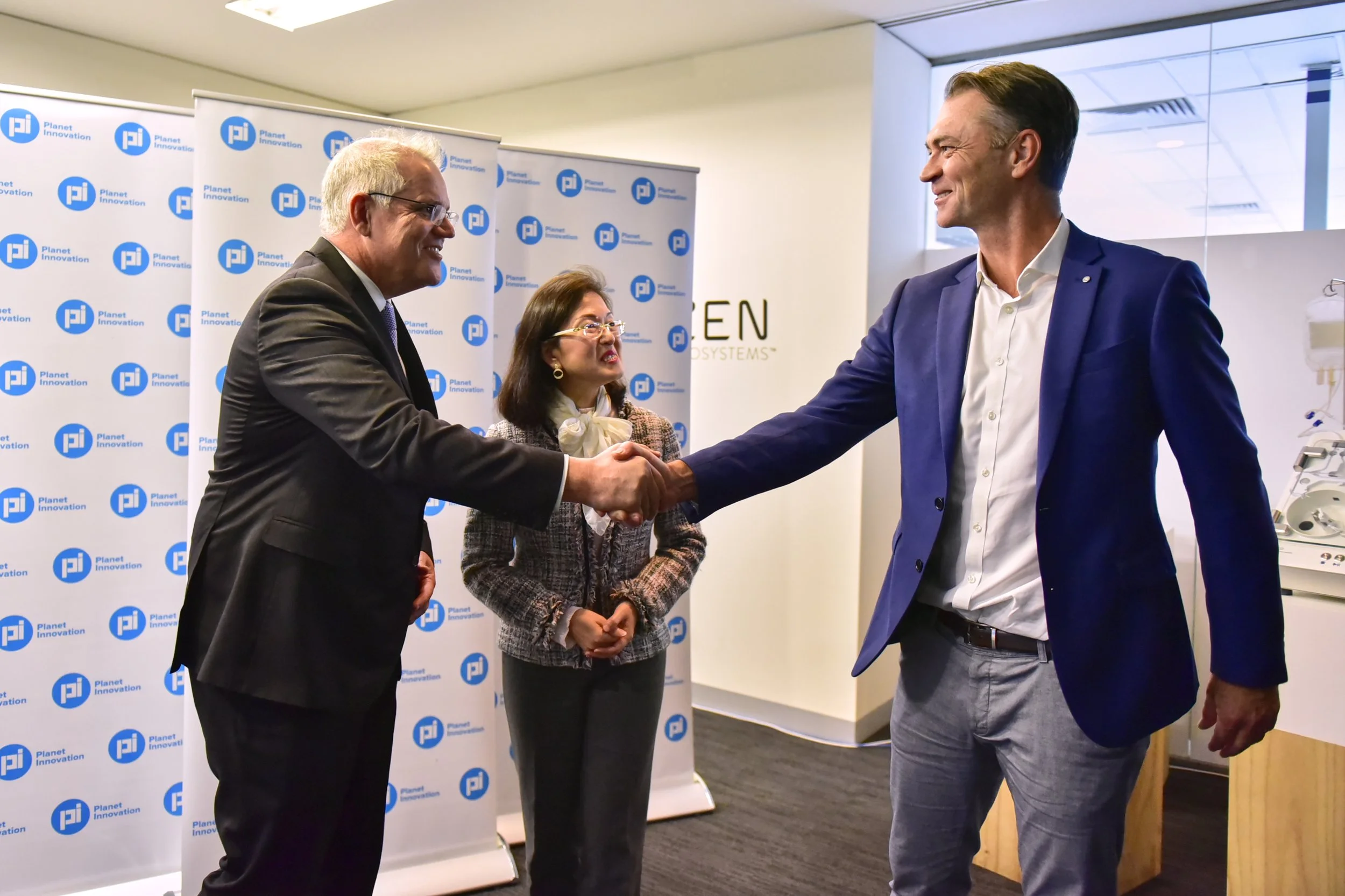 Two men shaking hands in a business setting, with a woman watching in the background. There are banners with the Planet Innovation logo behind them and a partial view of medical equipment on the right.