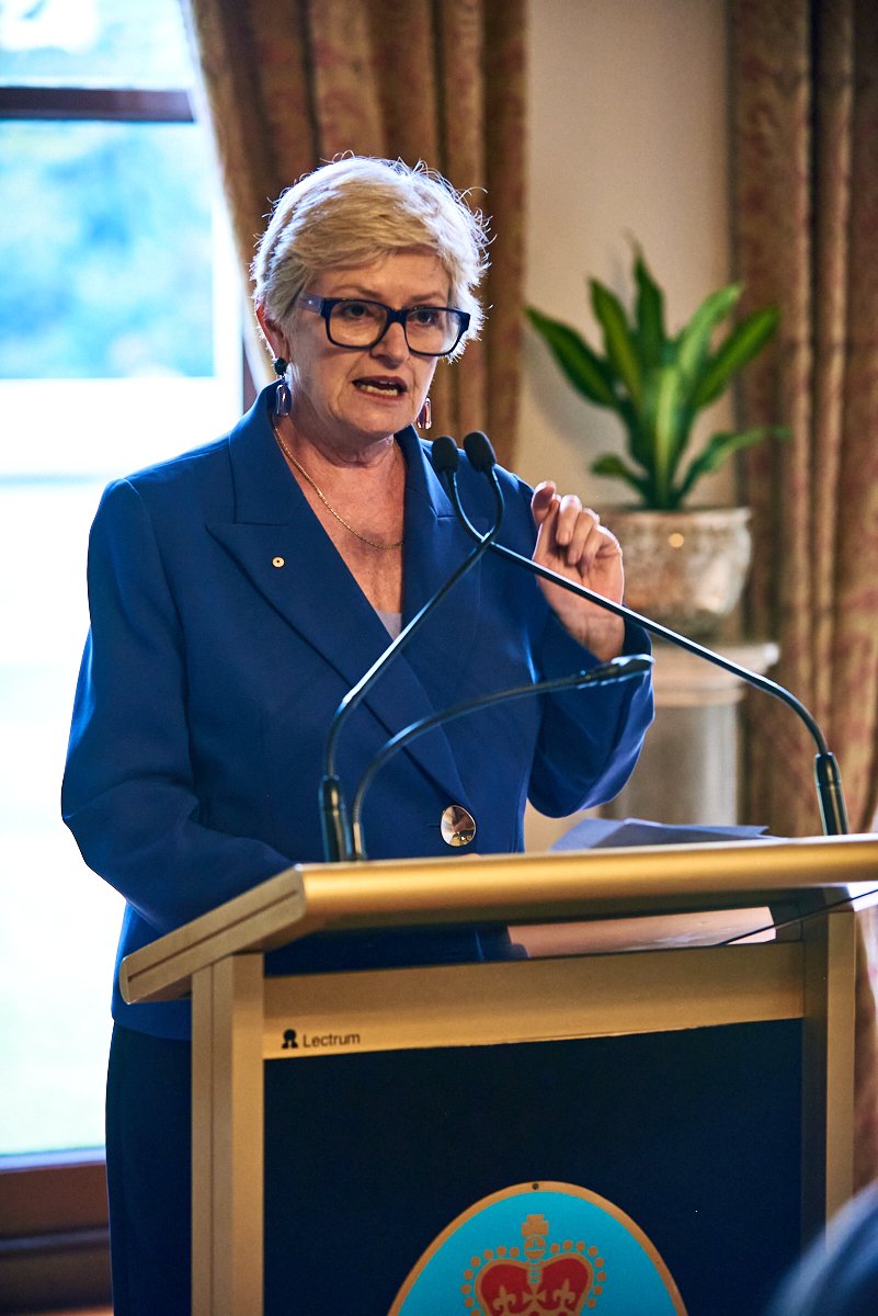 A woman with short blonde hair, wearing glasses and a blue blazer, speaking at a podium with a microphone in a room with a window and a potted plant in the background.