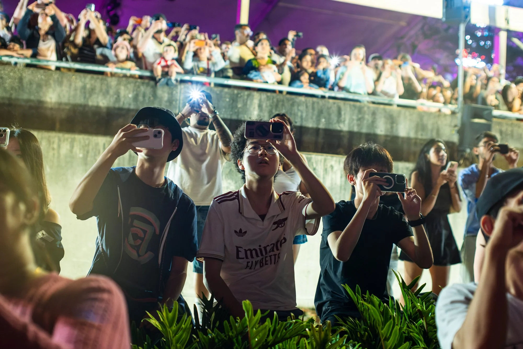 Crowd of people at an event, taking photos and videos with their phones, with purple lighting in the background.