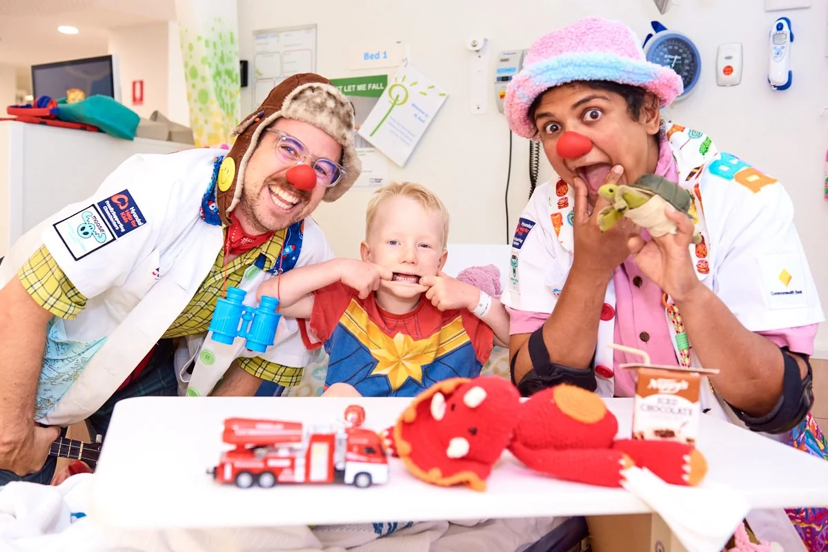 Two clown doctors and a young boy in a hospital room, dressed in colorful costumes with clown noses, making funny faces.