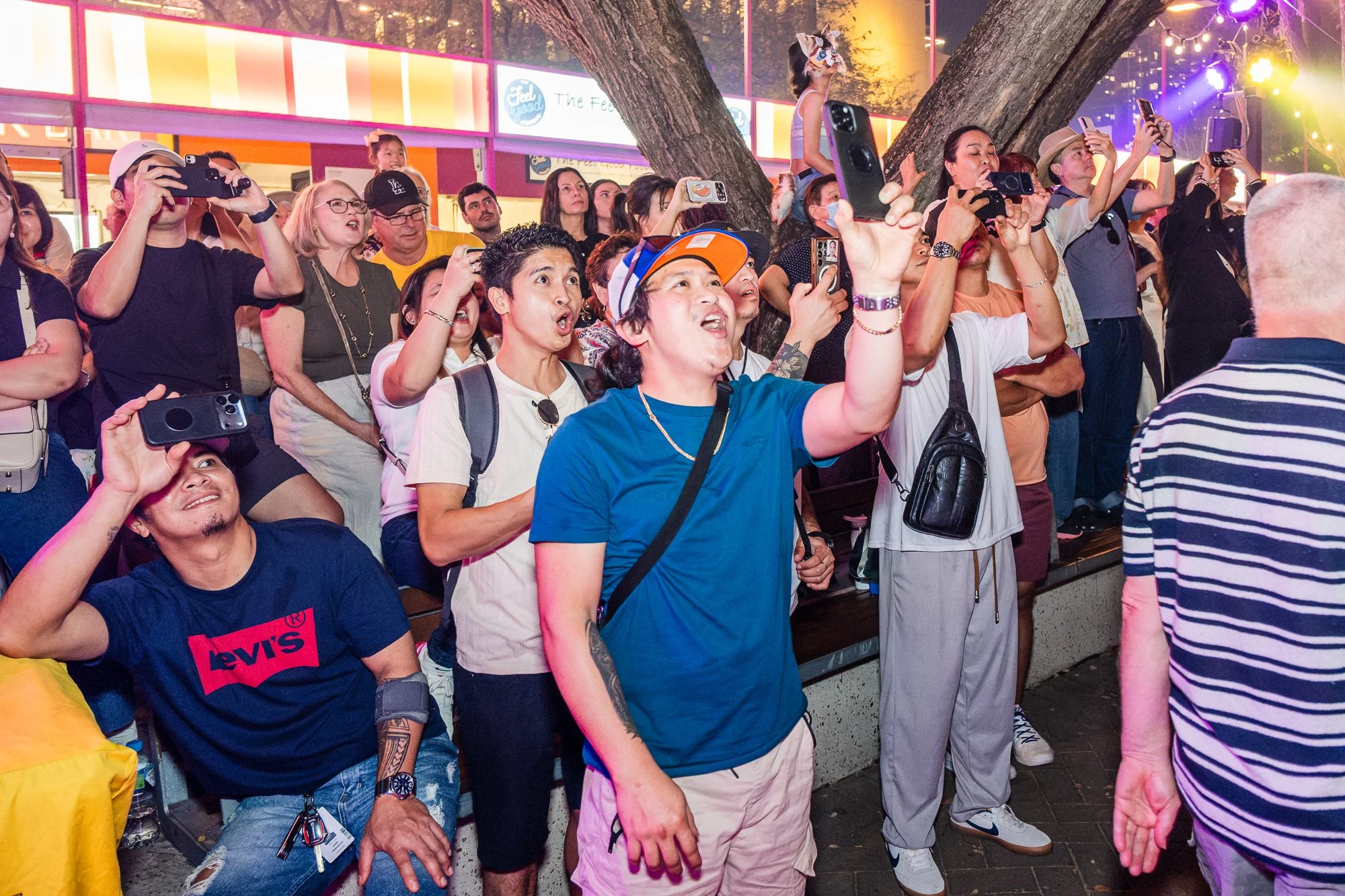 A crowd of people gathered outdoors at night, many of whom are taking photos or videos with their phones. The scene is brightly lit with colorful lights, and a large tree is prominent in the background. People exhibit gestures of excitement and atten