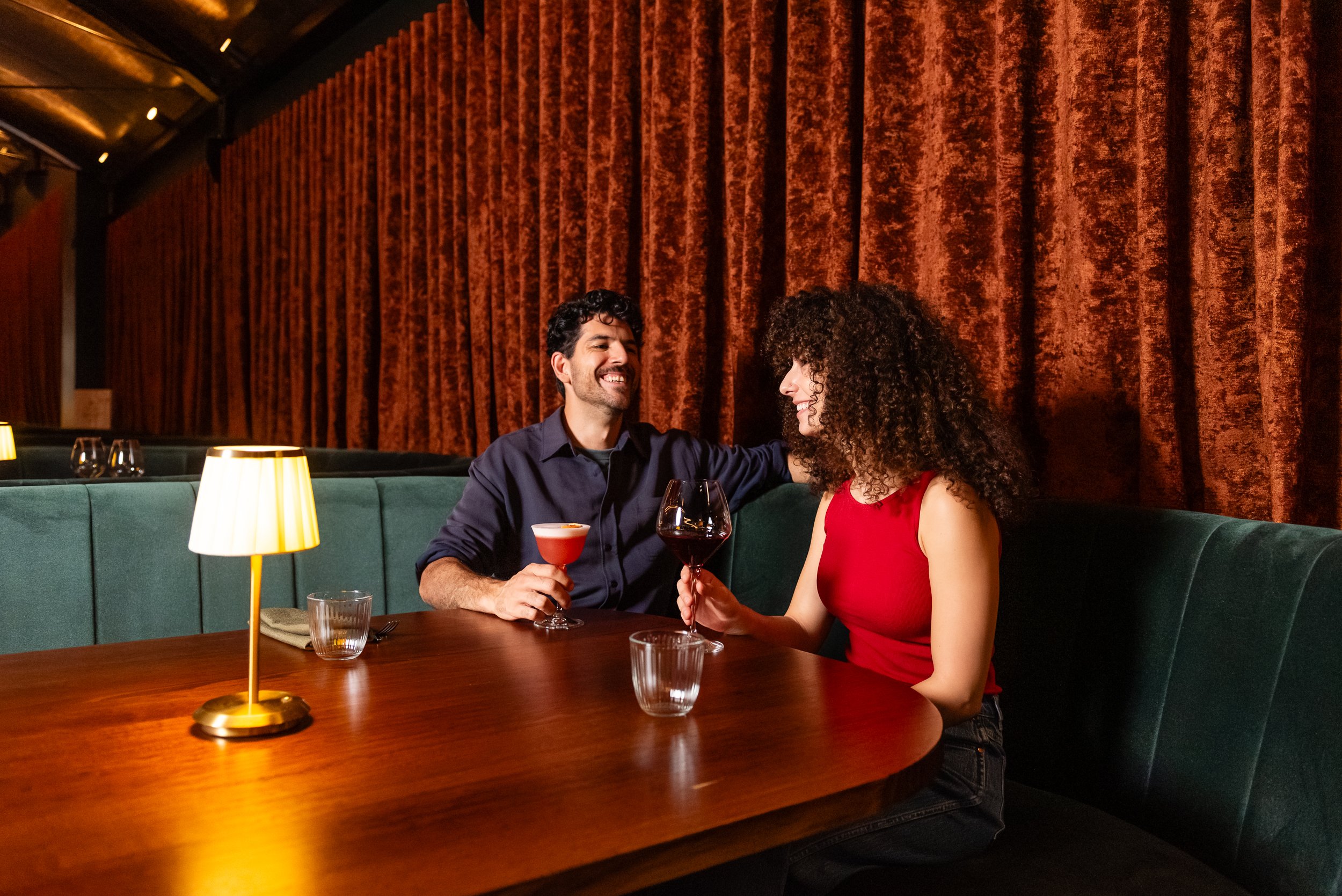 A man and a woman sitting at a restaurant table, enjoying drinks and smiling at each other. The man is holding an artfully garnished cocktail, while the woman has a glass of red wine. The background features rich, red velvet curtains, and a warm, amb