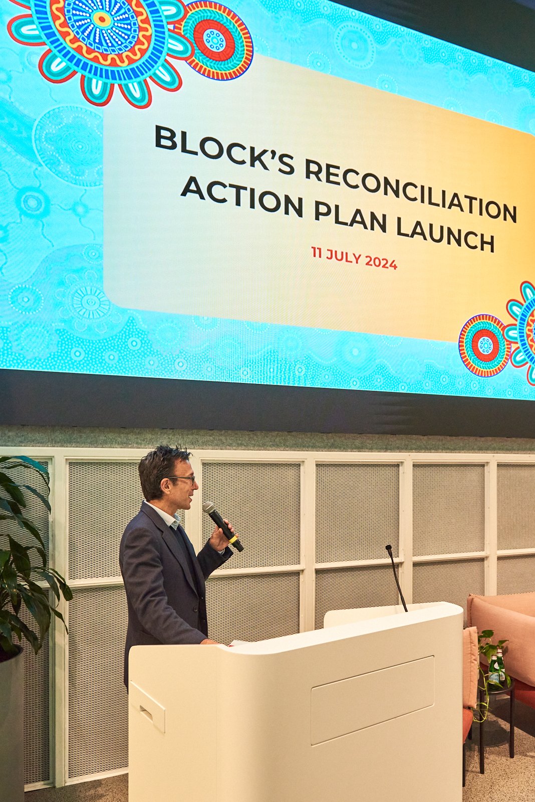 A man in a suit speaking into a microphone at a podium, with a large screen above him displaying 'BLOCK'S RECONCILIATION ACTION PLAN LAUNCH' and the date '11 JULY 2024'.