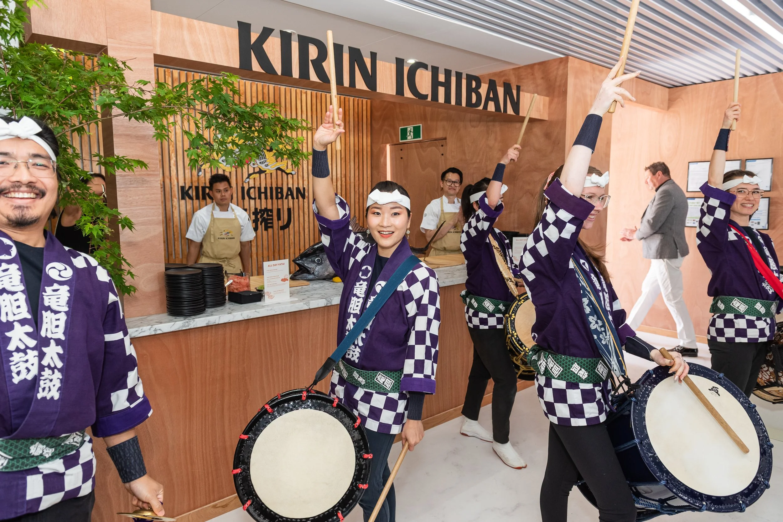 People dressed in traditional Japanese attire holding drums, performing a festival dance inside a restaurant with a sign that reads 'KIRIN ICHIBAN'.