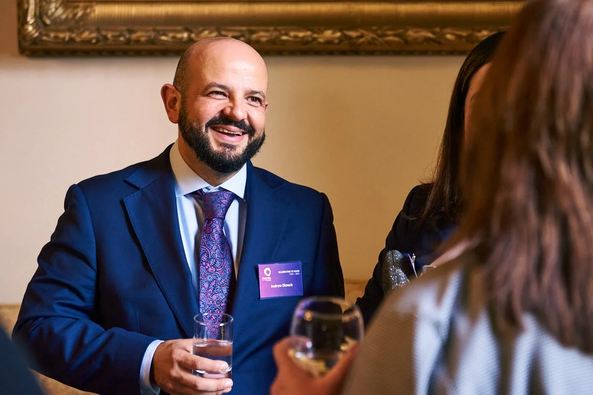 A man in a blue suit and patterned tie, holding a glass of water, is smiling and talking to women at a social event, in a room with beige walls and a framed picture.