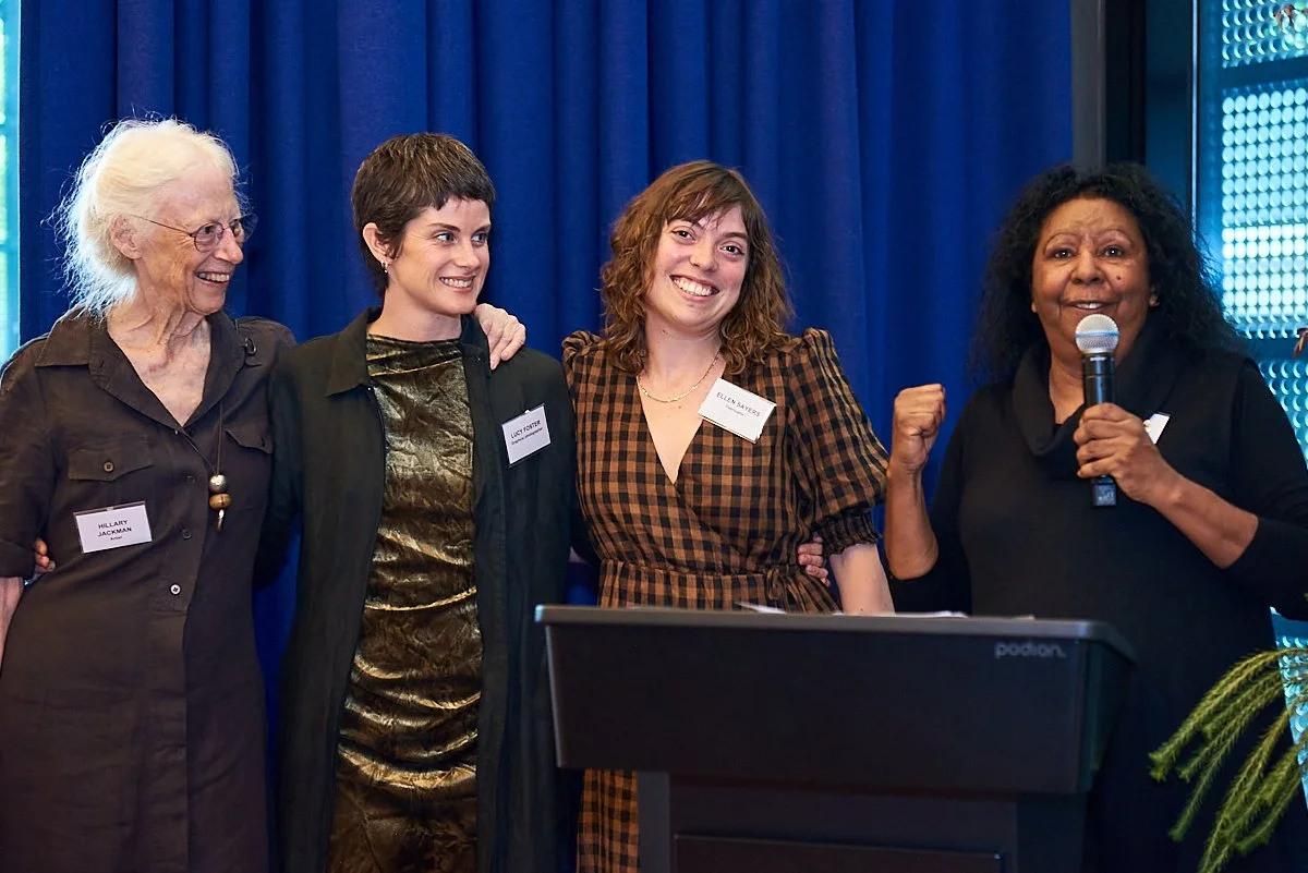 Four women standing together on a stage, smiling; one woman holding a microphone, with a blue curtain in the background.