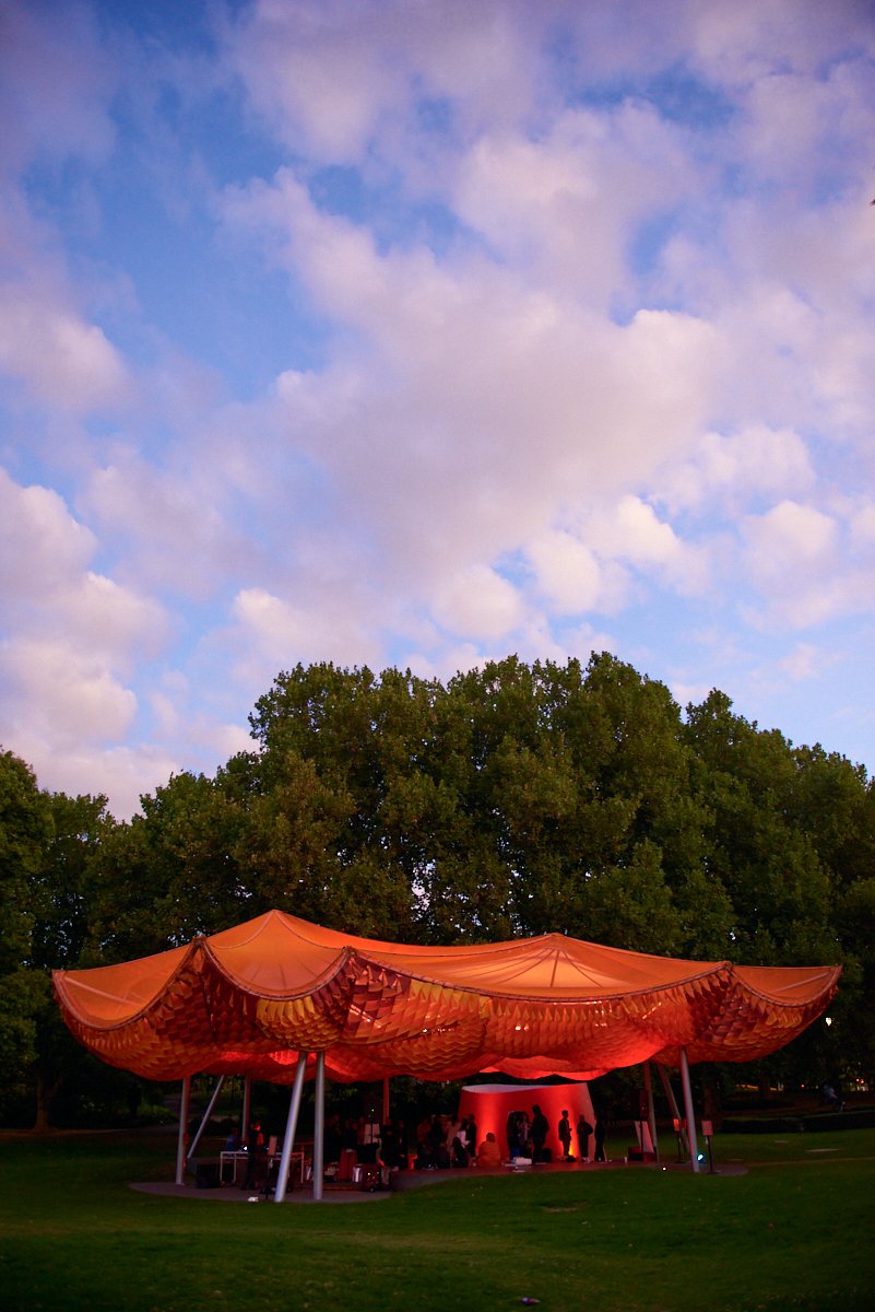 An outdoor event under a large, orange, canopy-like structure with people gathered underneath, set in a park with trees and a blue sky with clouds.