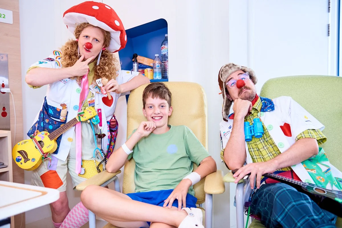 Two clowns and a boy in a hospital room, with the boy smiling while seated in a chair, and the clowns striking playful poses.
