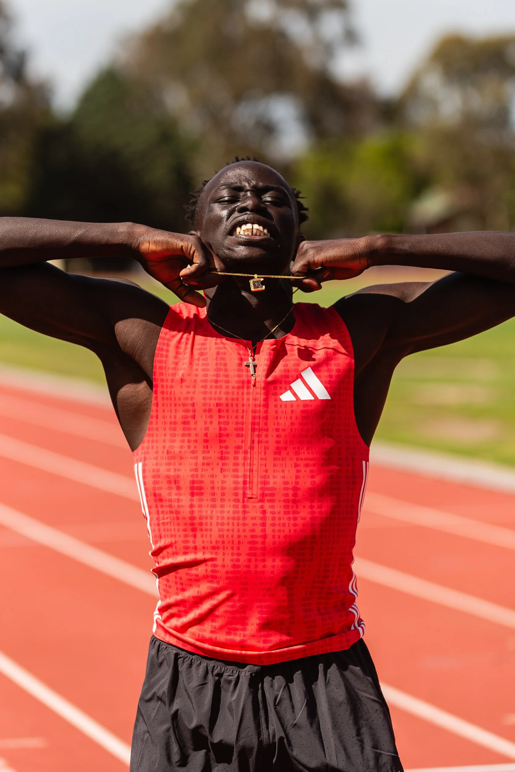 A male sprinter in a red athletic outfit, stretching his neck with his hands, at a running track outdoors.