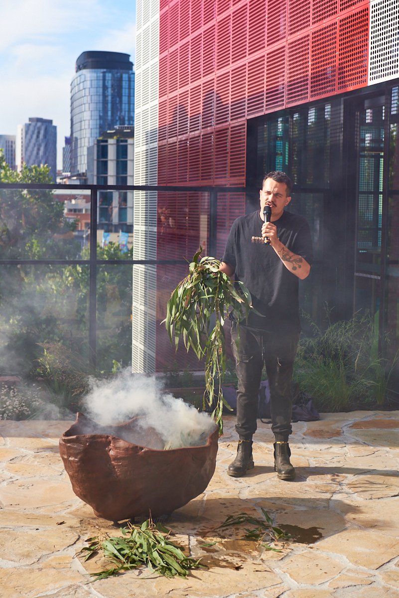 A man in black clothing holding a microphone and a leafy plant, standing near a large pot with steam rising from it on a rooftop with city buildings in the background.