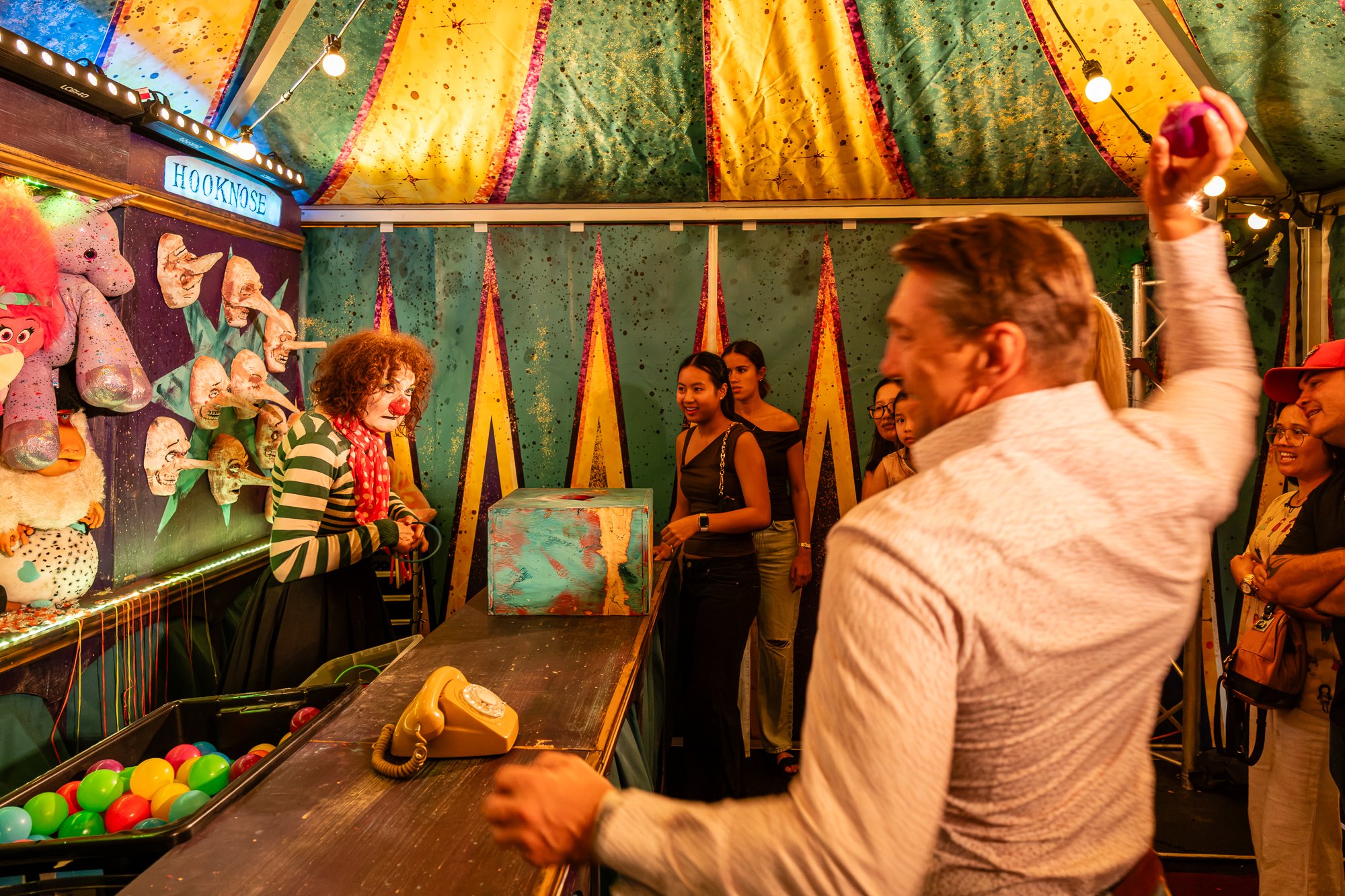 People at a carnival game booth with a colorful, starry backdrop, a clown with a red nose and striped shirt, and a variety of plush toys and game balls.