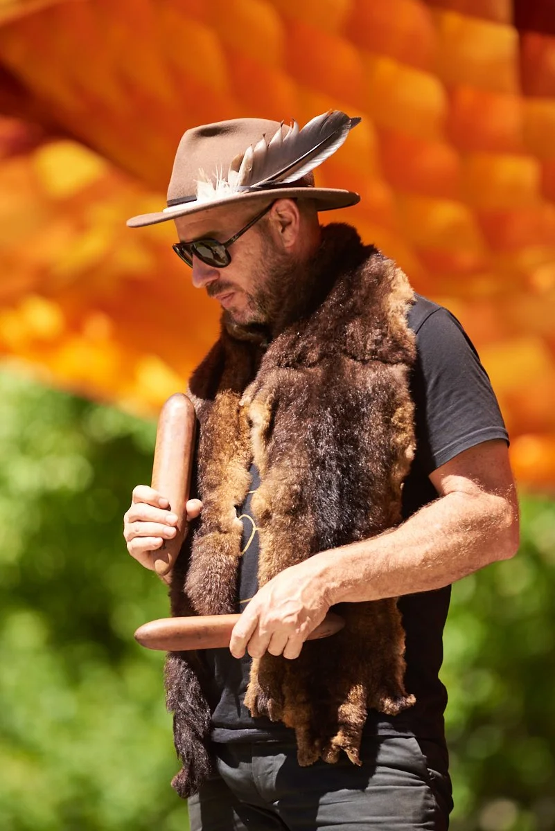 Man wearing sunglasses, a fur scarf, and a brown hat with a feather, holding wooden sticks, outdoors with blurred vibrant orange, green, and yellow foliage in the background.