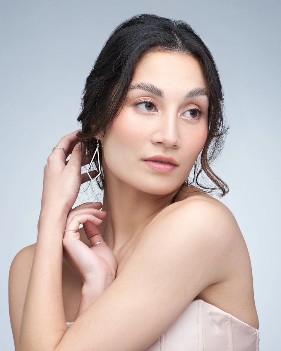 A young woman with dark hair, wearing earrings, and a strapless light-colored top, looking to the side with a soft expression.