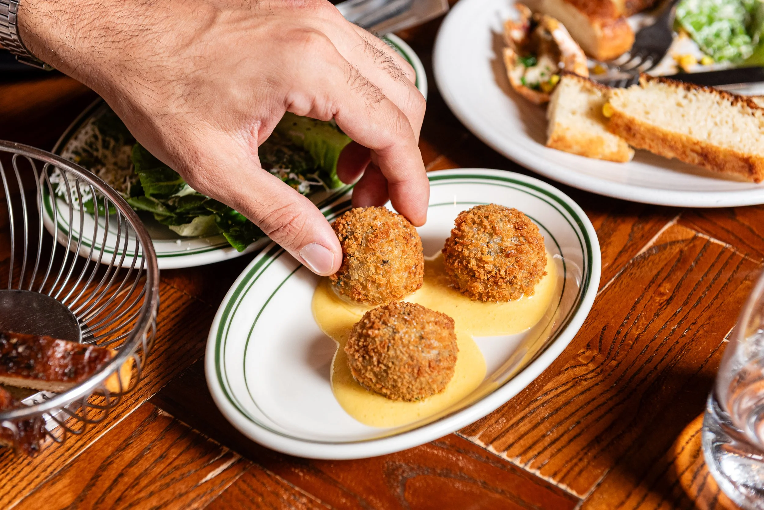 A hand placing breaded croquettes on a white plate filled with mustard sauce, with a side salad, baked potato, and additional food items on plates in the background.