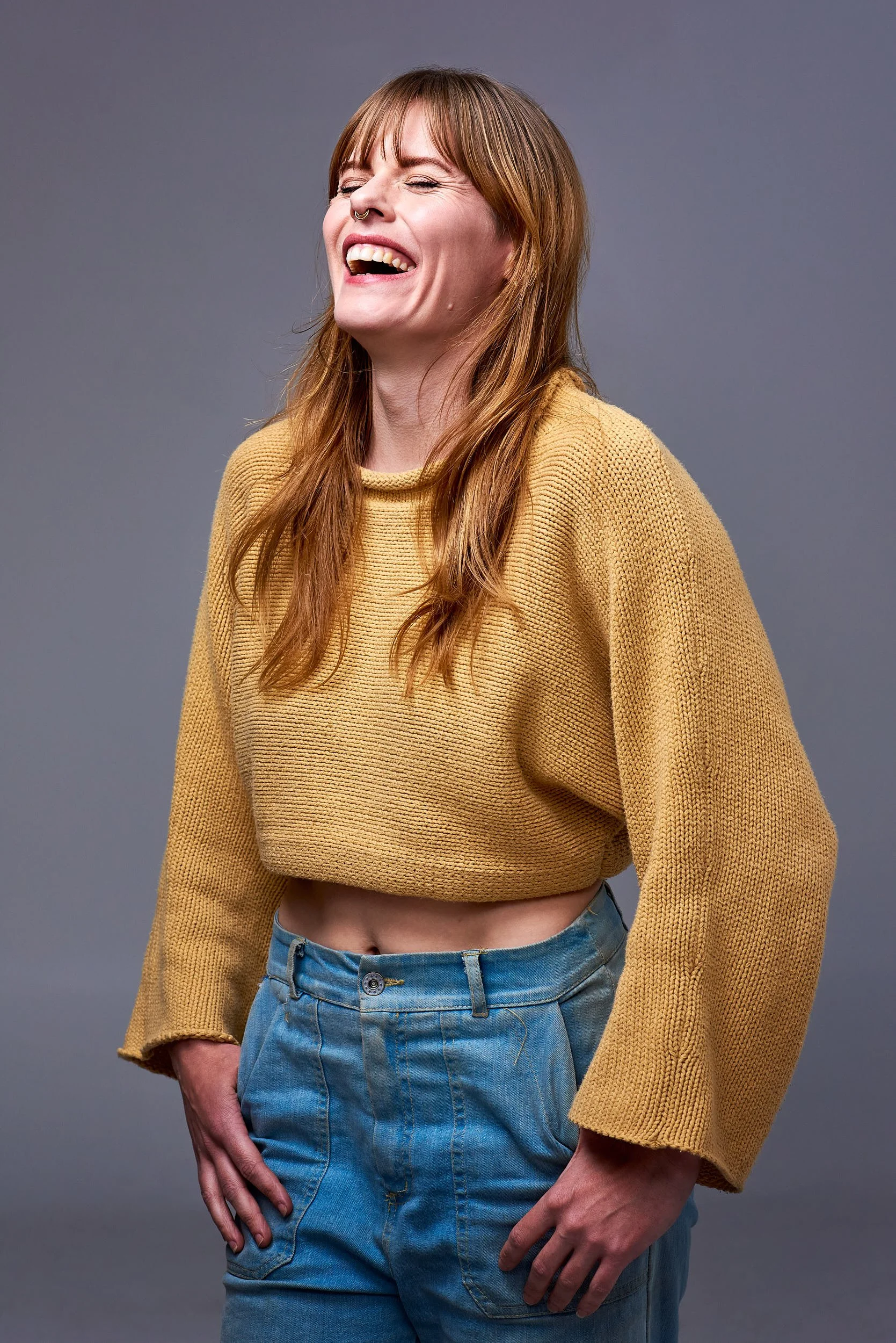 A woman with red hair and freckles, wearing a yellow cropped sweater and high-waisted jeans, laughing with eyes closed against a gray background.