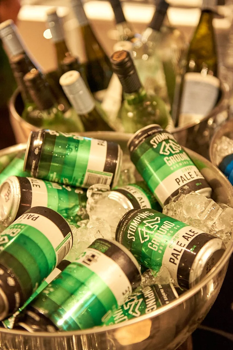 A metal bucket filled with ice and multiple cans of pale ale beer, with several green and black labels visible, and some beer bottles standing in the background.