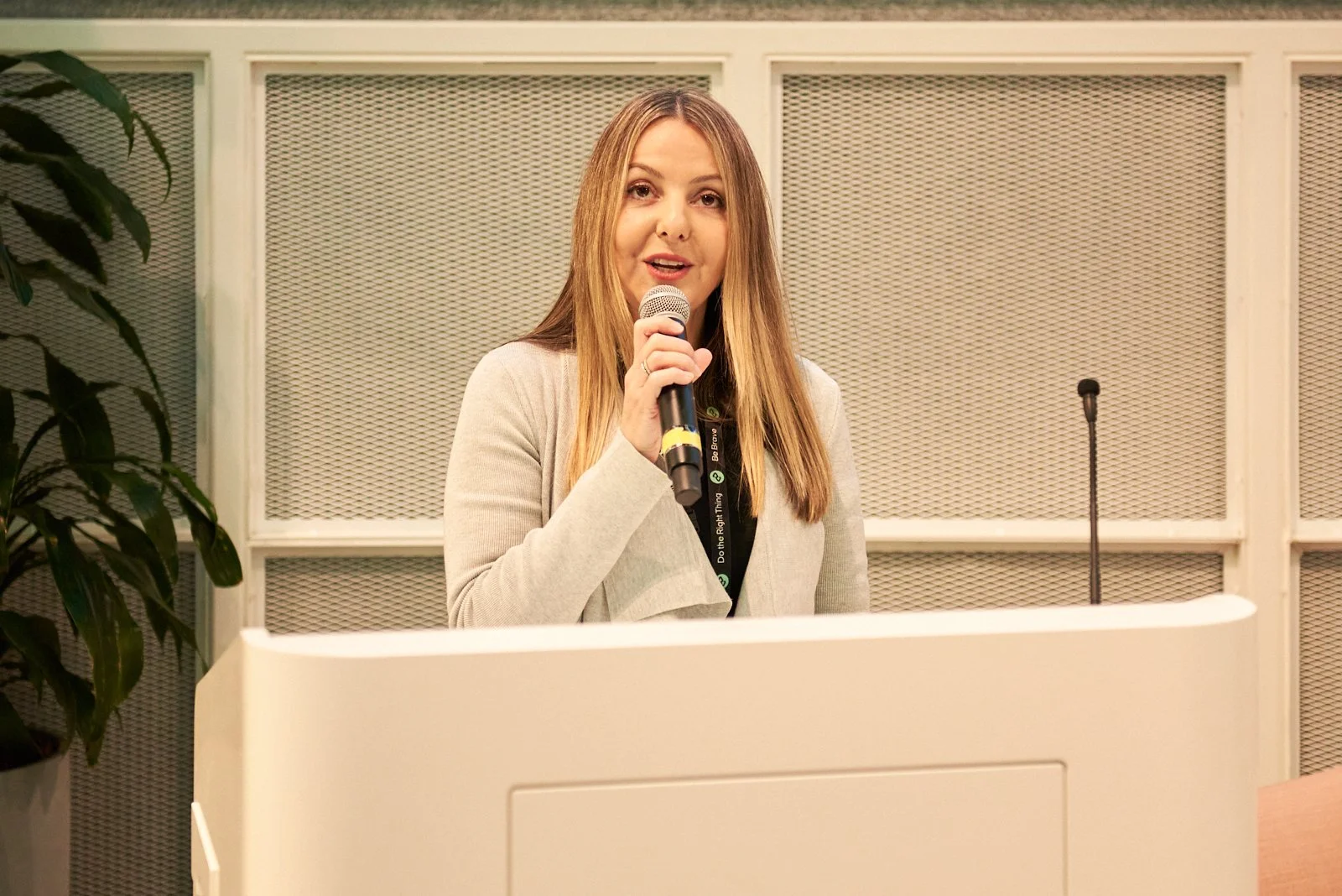 A woman with long light brown hair holding a microphone, standing behind a white podium in front of a metal mesh backdrop, with a potted plant on the side.