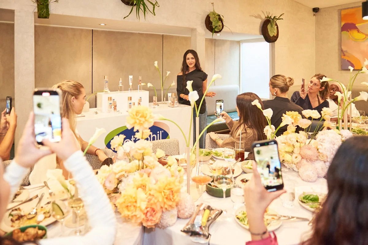 A woman giving a presentation at a table surrounded by women taking photos with their smartphones, with floral centerpieces and skincare products displayed behind her.