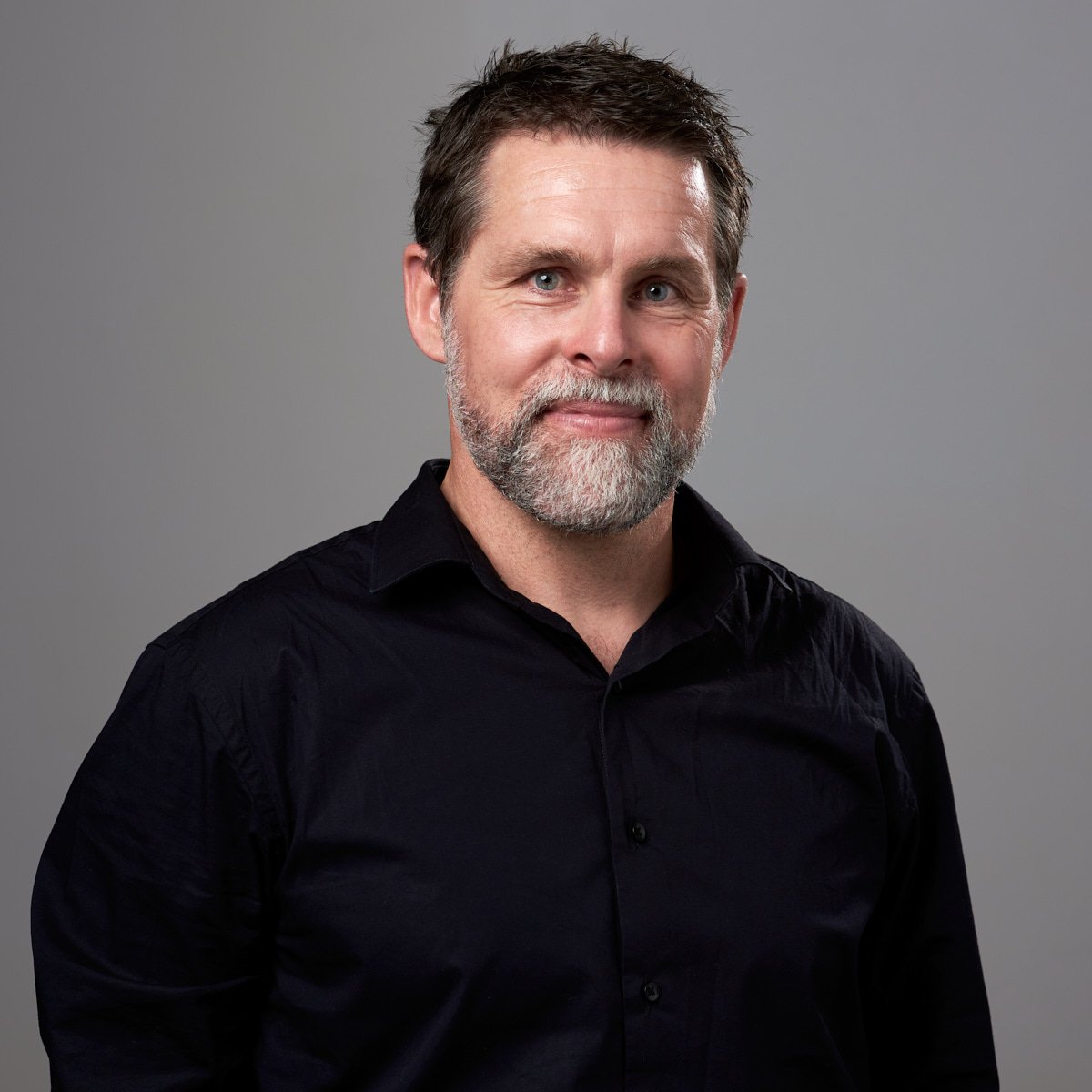 A middle-aged man with short brown hair, a gray beard, and blue eyes wearing a black button-up shirt against a gray background.