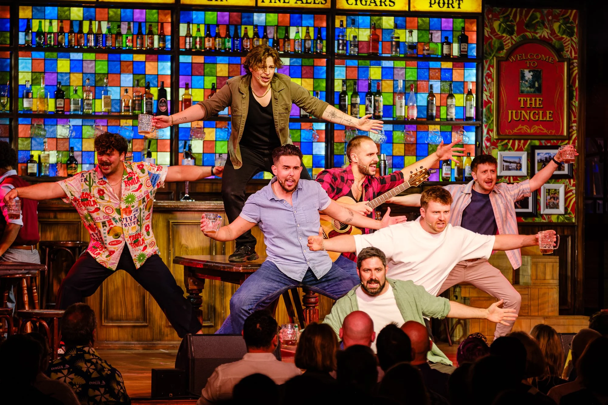 A lively scene in a bar or pub with multiple people performing on stage, singing, playing guitar, and dancing. The background features colorful stained glass shelves with various bottles of alcohol and a sign that reads 'The Jungle'. Audience members