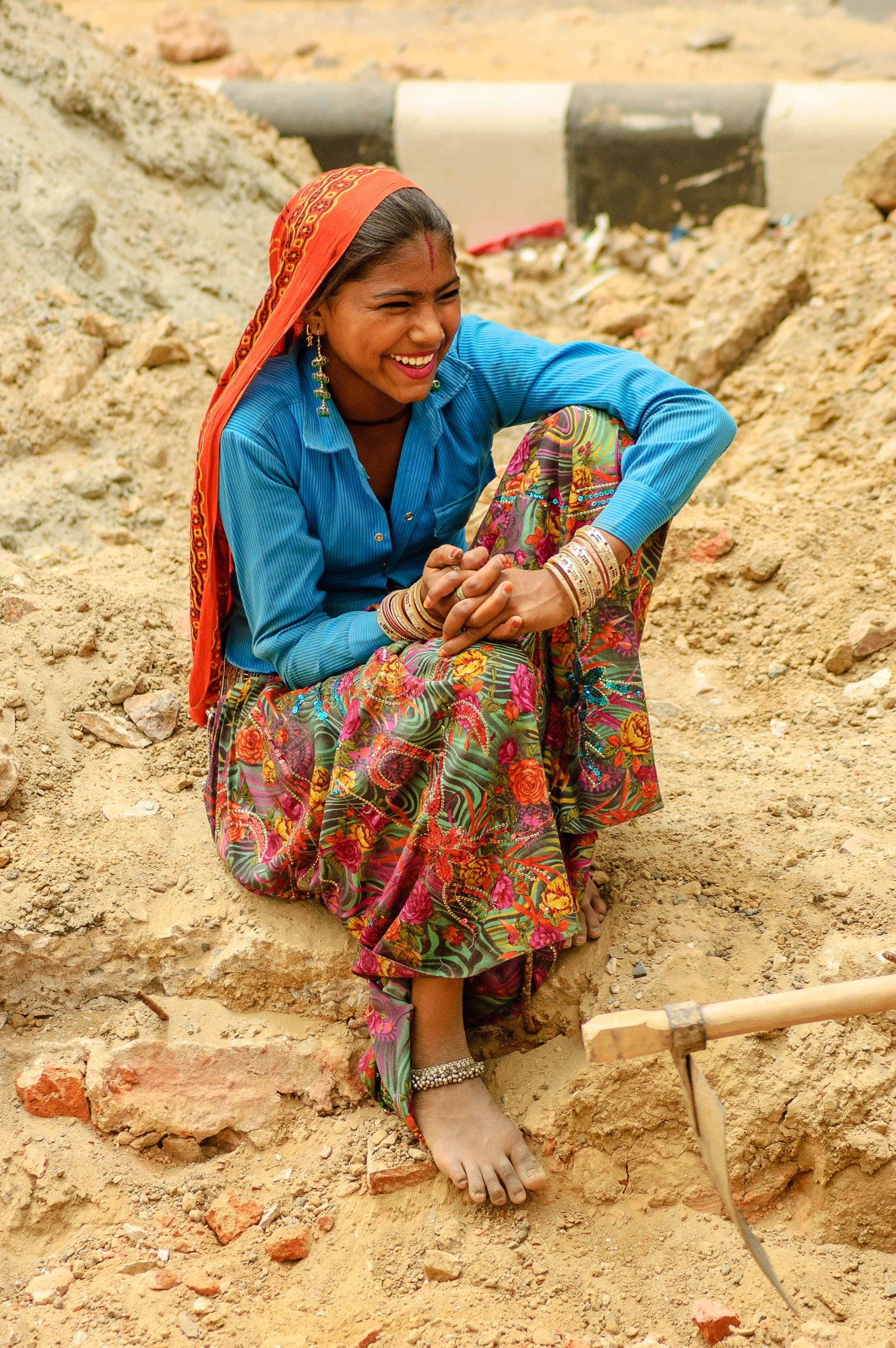 A woman sitting on a sandy area, smiling and wearing colorful traditional clothing with jewelry, with a specter of construction or excavation work in the background.