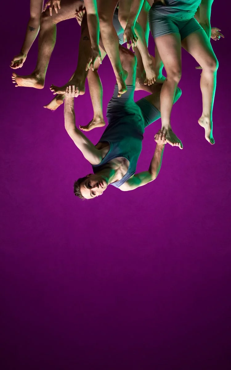 A group of seven people holding each other upside down against a purple background, with only their legs and one person's face visible.