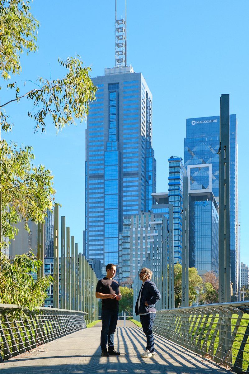 Two men having a conversation on a bridge in an urban park with modern skyscrapers in the background against a clear blue sky.