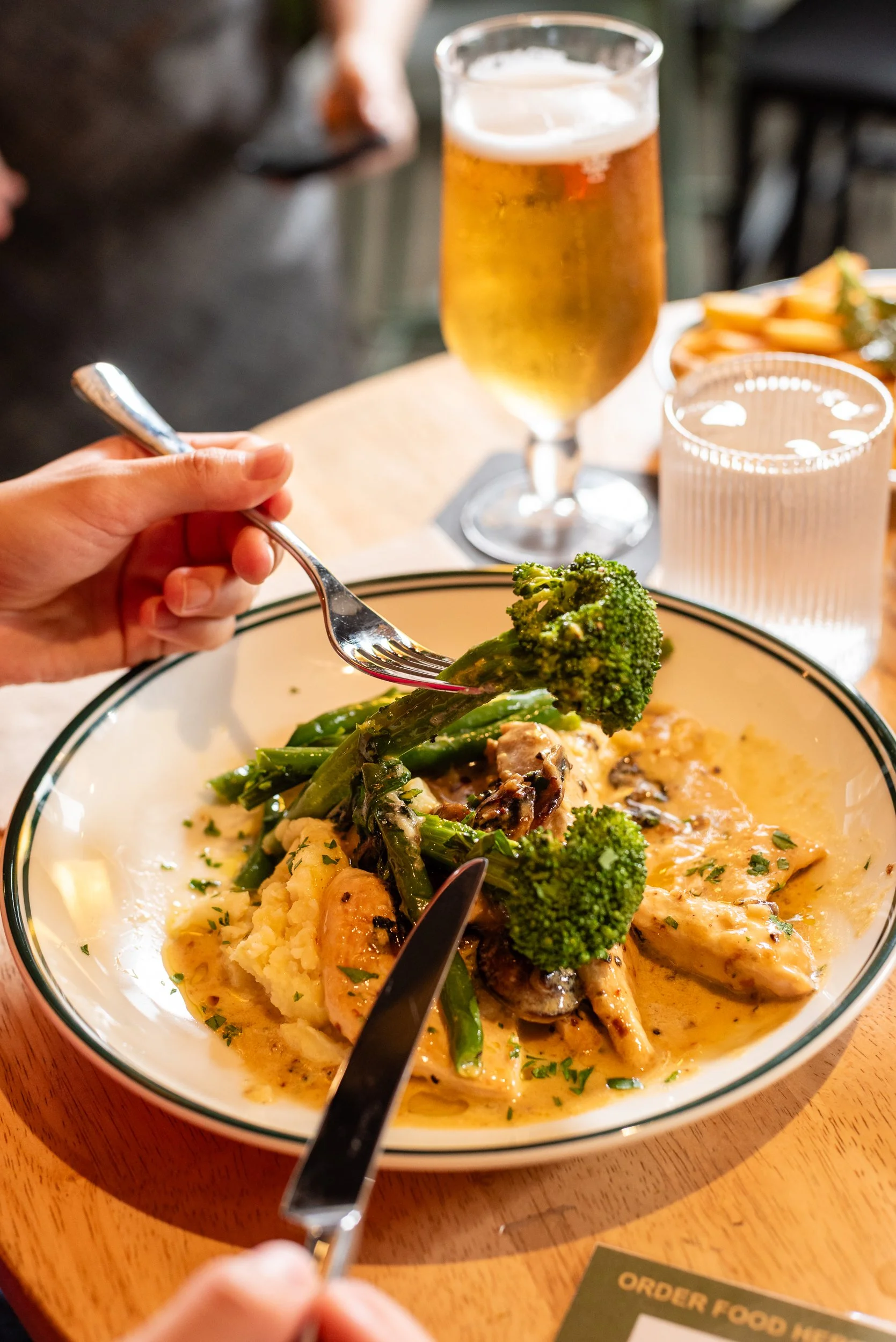 A bowl of food with mashed potatoes, green broccoli, and chicken in sauce. A person is holding a fork with broccoli. In the background, there is a glass of beer and a glass of water with ice. The setting appears to be a restaurant or dining table.
