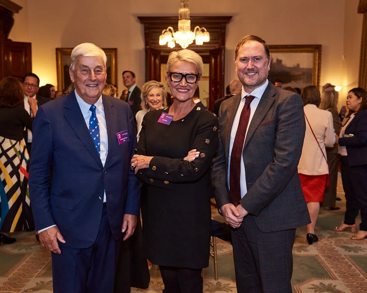Three well-dressed individuals, two men and one woman, standing together in a formal setting at a social or professional event. The woman is in the center, smiling with crossed arms, while the men stand on either side of her, smiling. The room has wa