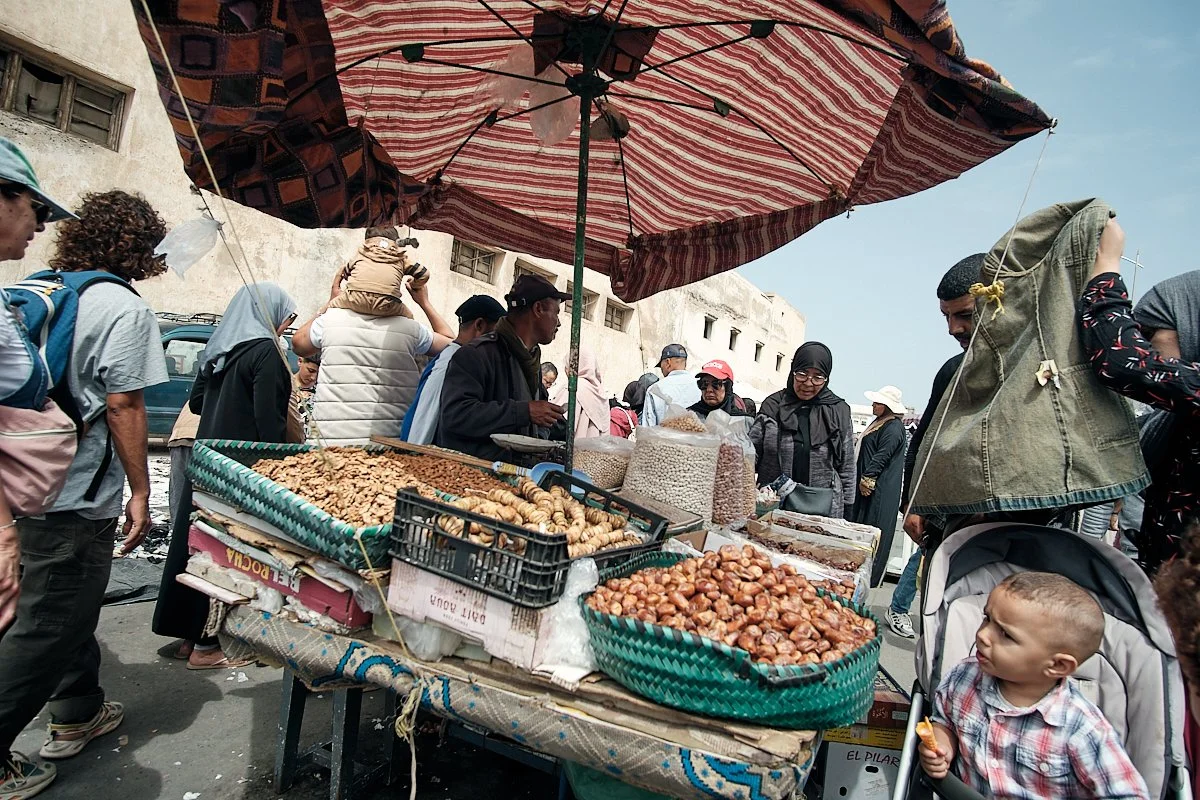Street market scene with a vendor stall selling nuts and snacks, people shopping and walking around, an umbrella shading the stall, and a building with small windows in the background.