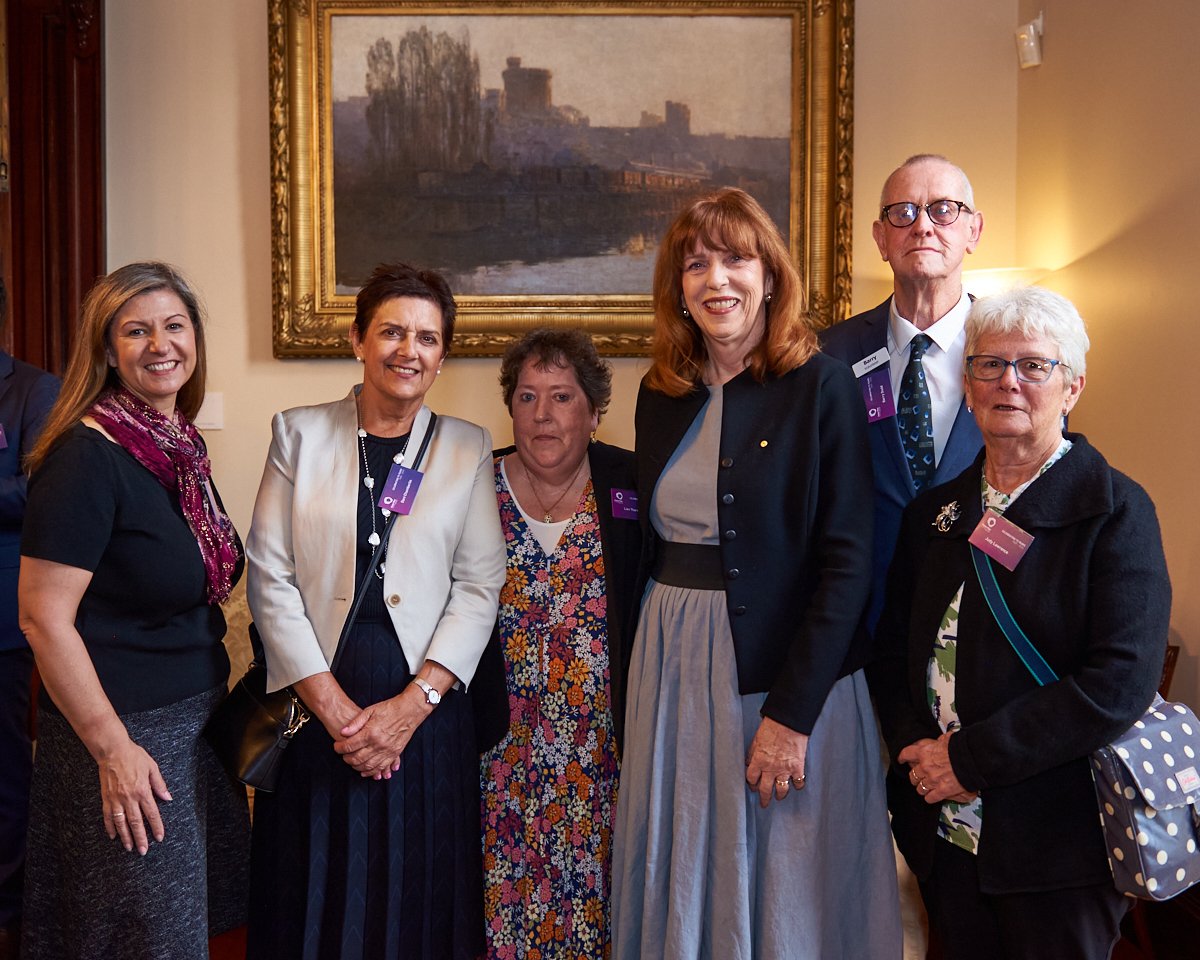 Group of six women and one man standing together indoors at a formal event, smiling, with a framed painting on the wall behind them.