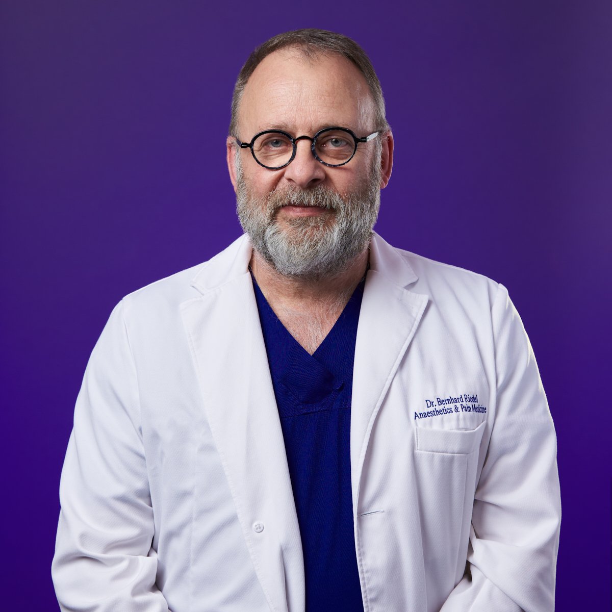 A doctor with glasses, a gray beard, and wearing a white coat standing against a purple background.