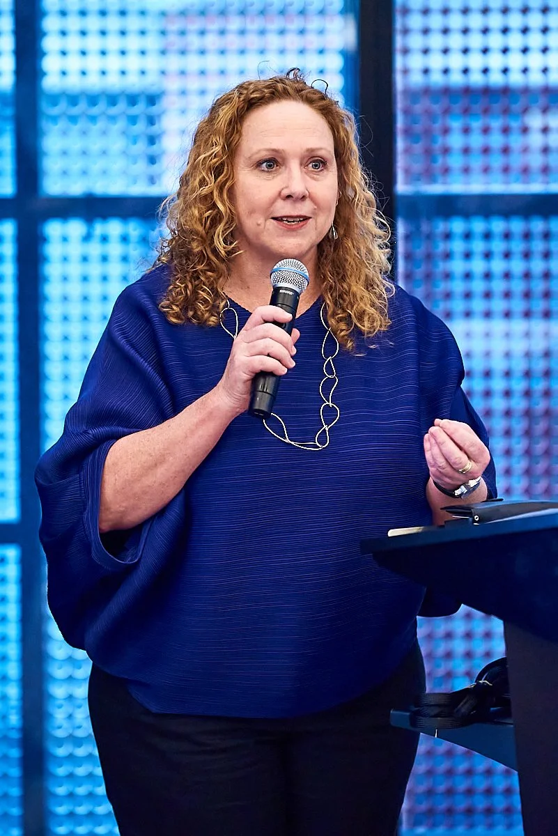 A woman with curly red hair, wearing a blue top and a silver necklace, holding a microphone and speaking at a presentation or event.