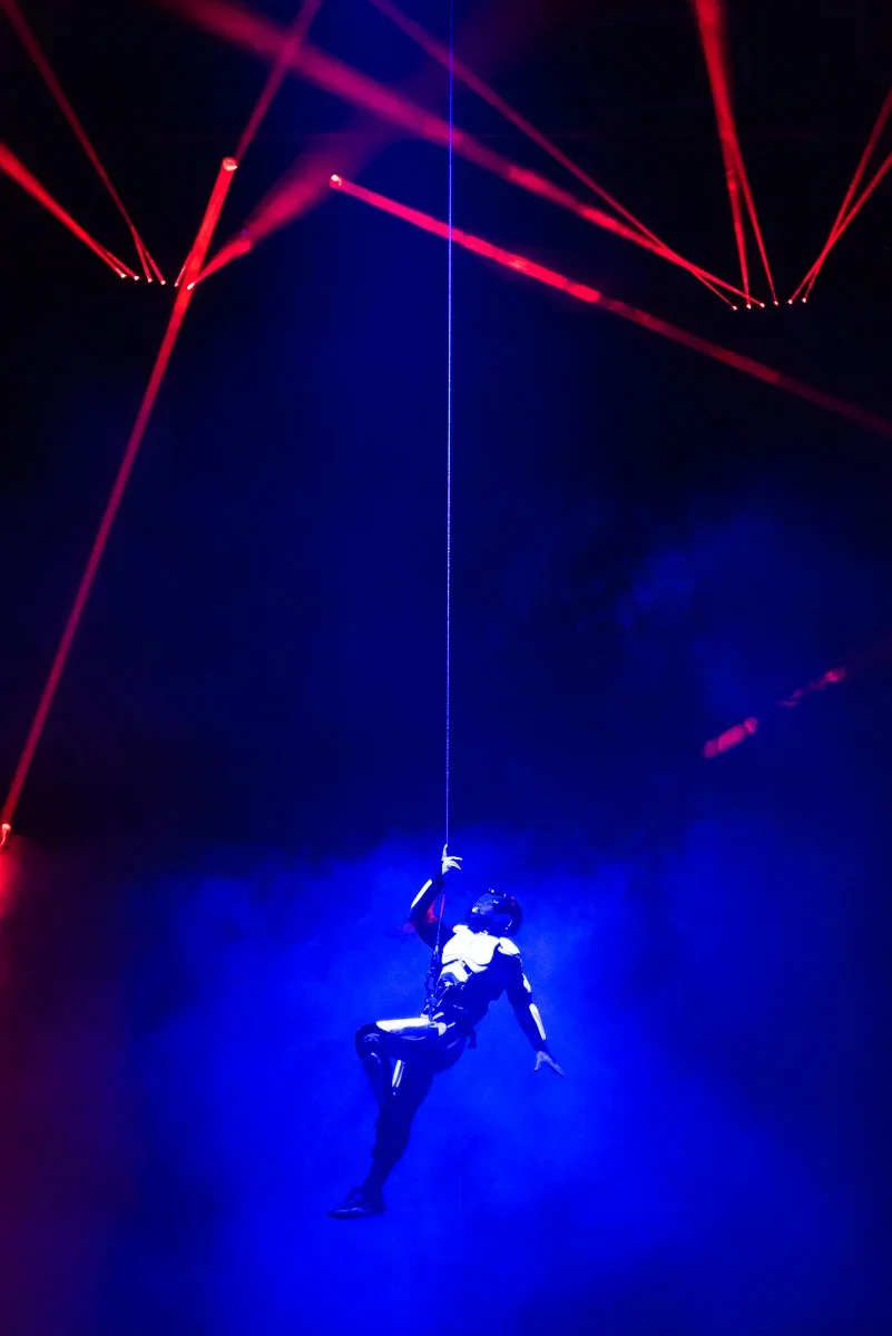 An acrobat performing suspended from a rope or cable with blue lighting and red laser light beams in a dark setting.