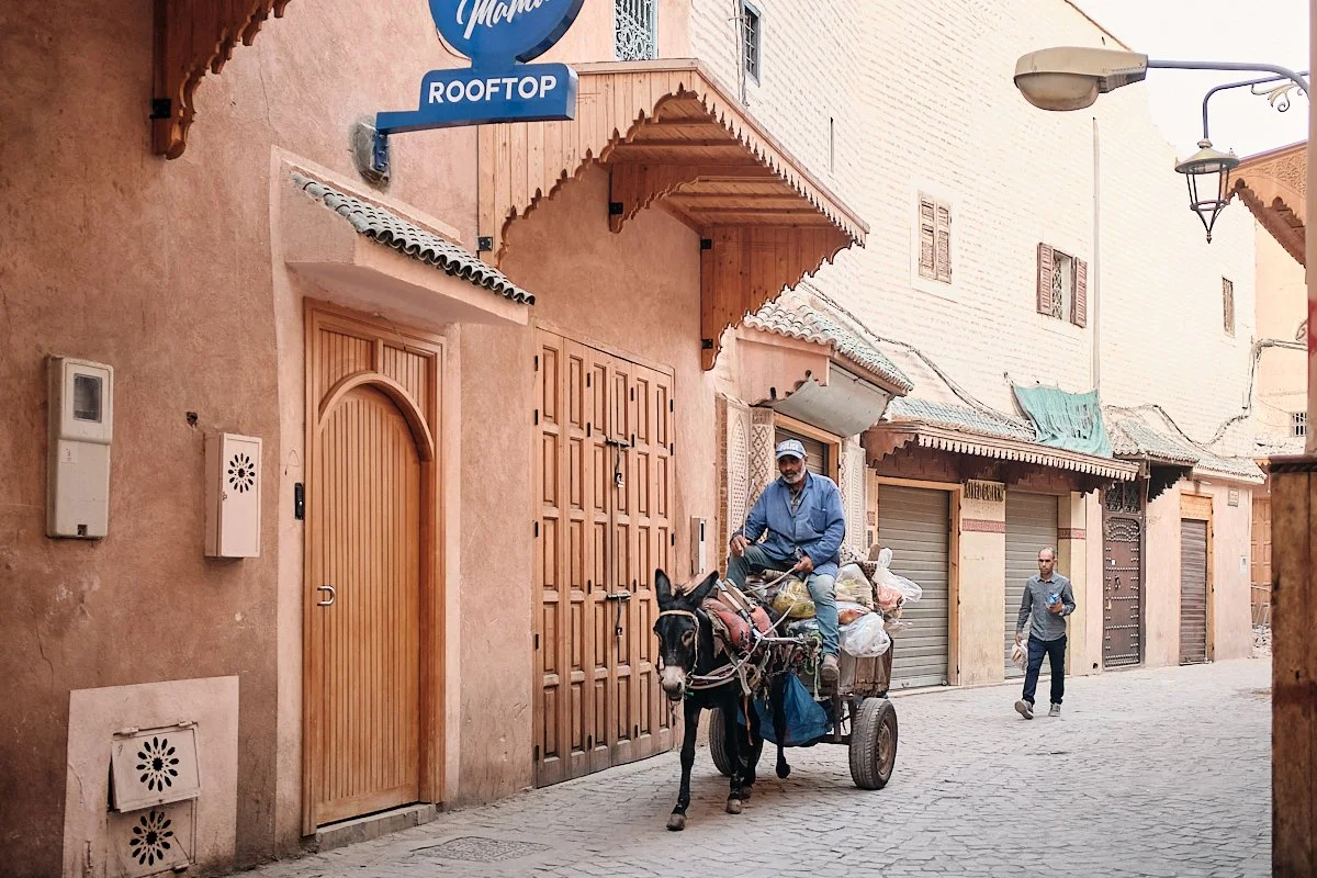 A man riding a donkey that is pulling a cart loaded with bags down a narrow street with closed shops and buildings with wooden doors and shutters, in an urban area.