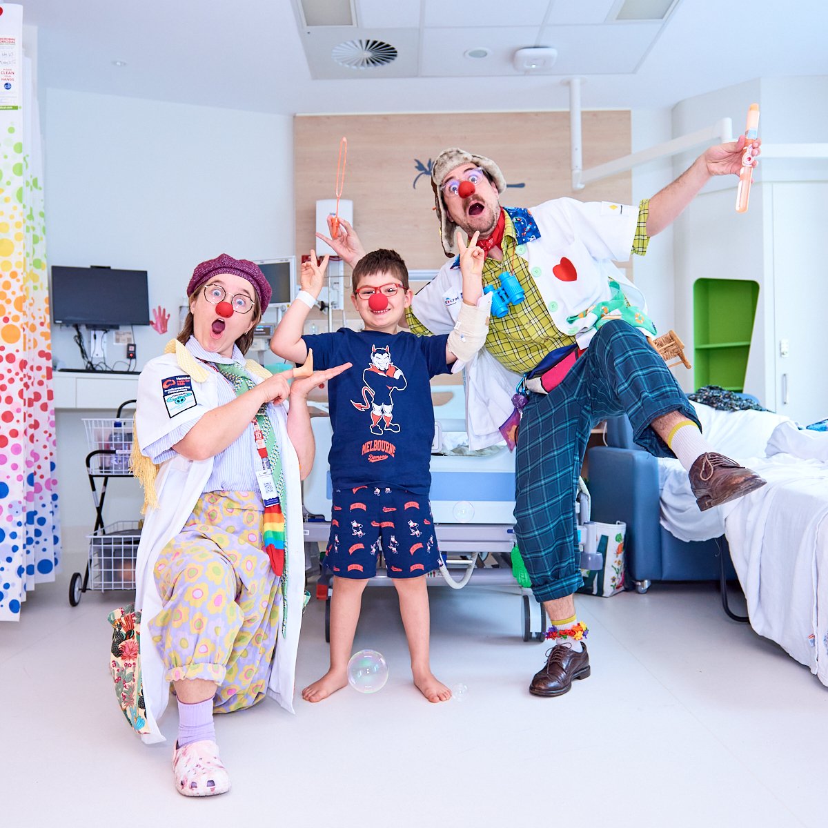 Three people dressed as clowns in a hospital room, smiling and making playful gestures. One woman is kneeling with painted face, glasses, and a colorful outfit. A boy with a red nose and glasses is standing in the middle. An older man with a clown no