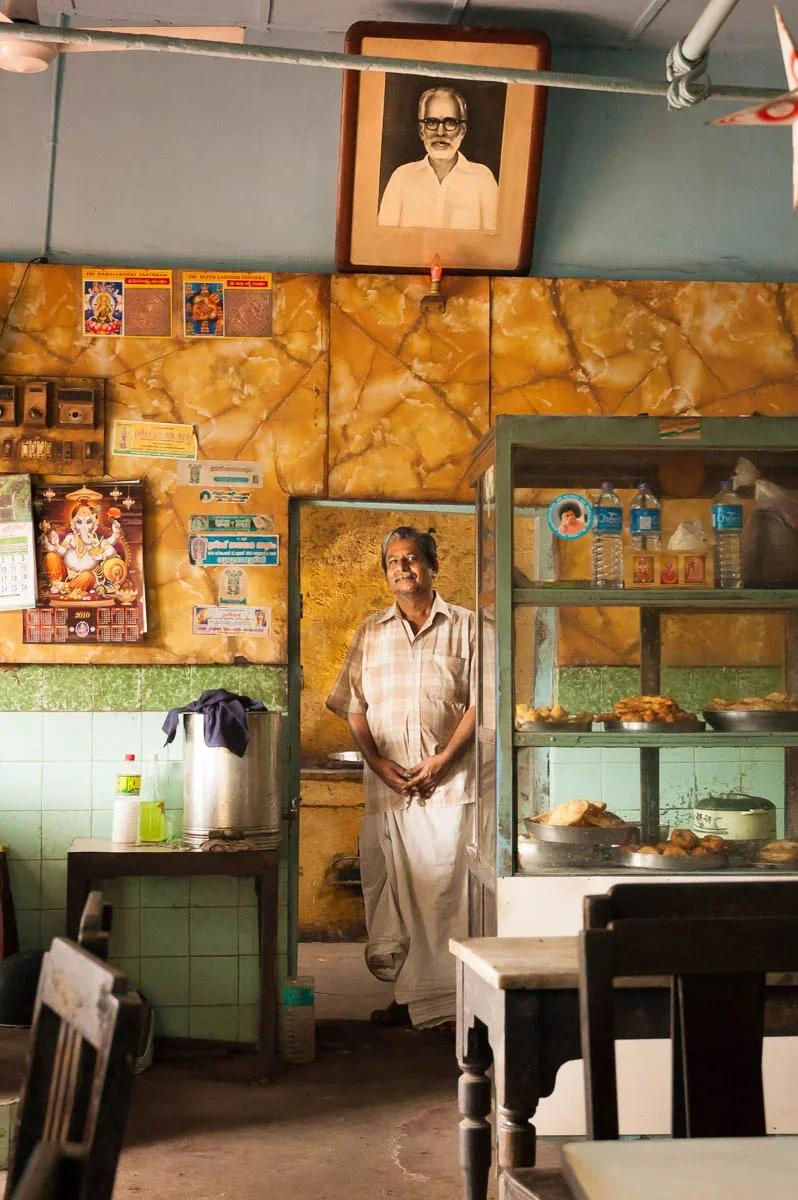 A man standing inside a small eatery or shop, smiling at the camera. Behind him, there is a yellow marble textured wall, a framed portrait of a man with glasses hanging above. To the right, a glass display case with food items and bottled water. On the left, a calendar and some posters or notes on the wall. The interior has a mixture of green tiles and wooden furniture.