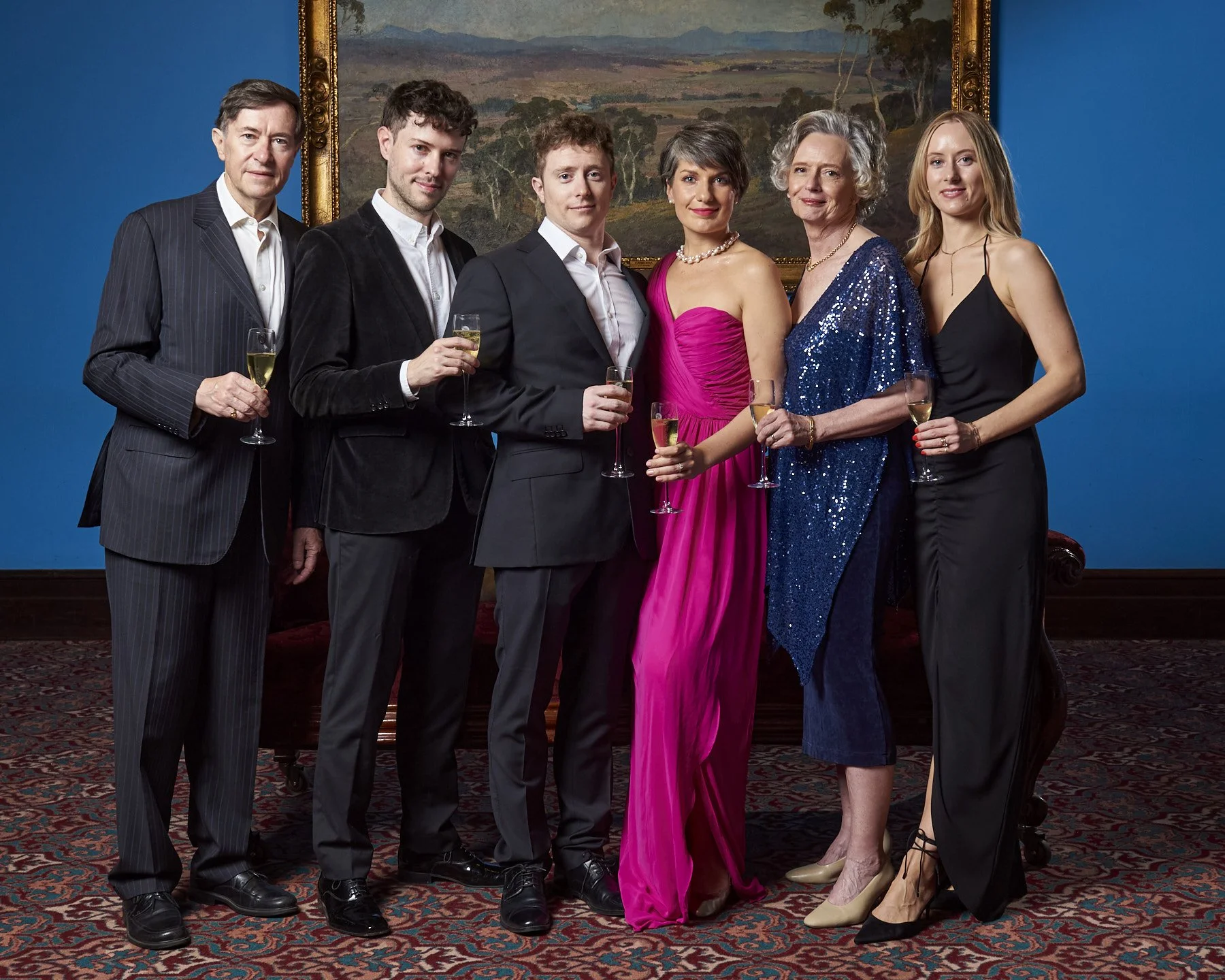 Seven people dressed in evening attire stand together in a formal setting, holding glasses of champagne. The group consists of four men and three women, with a large landscape painting hanging on a blue wall behind them.