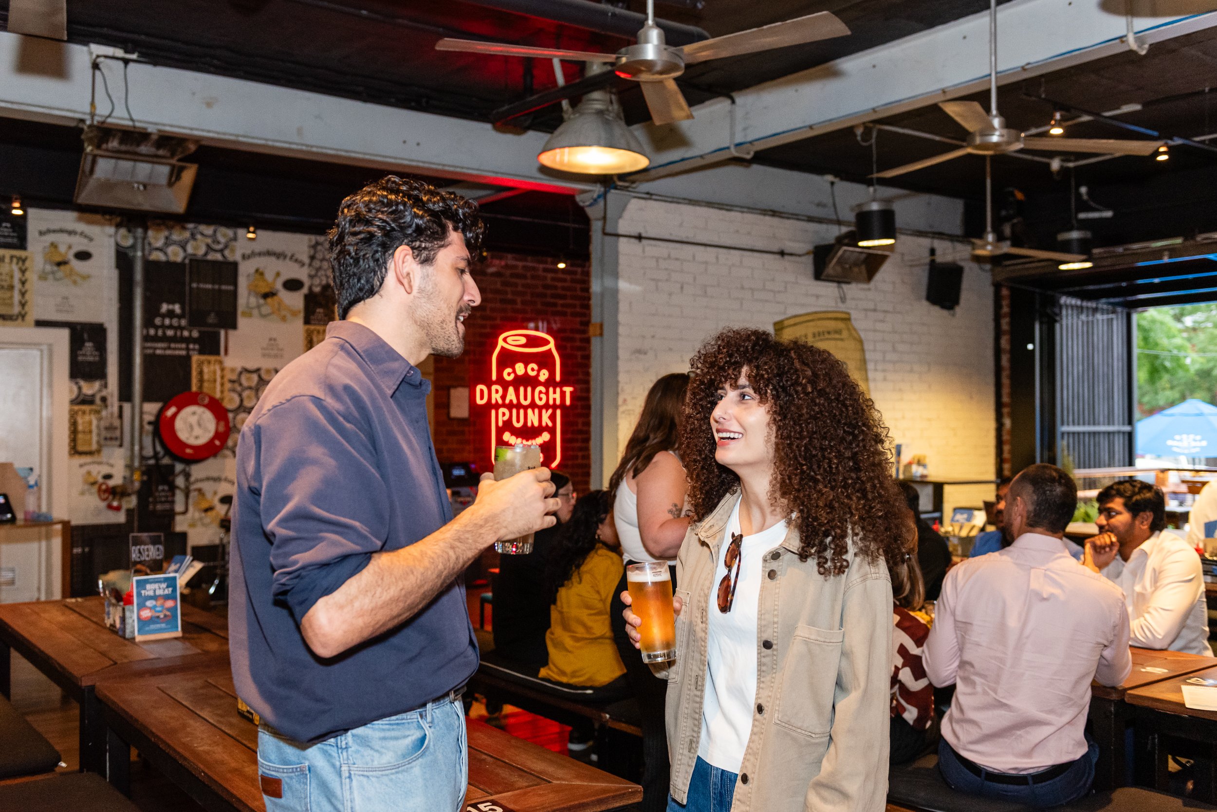 Two people, a man and a woman, are talking and smiling in a crowded bar or restaurant. The man is wearing a blue shirt and holding a drink, and the woman has curly hair and is also holding a drink. Behind them are other patrons and a neon sign that r